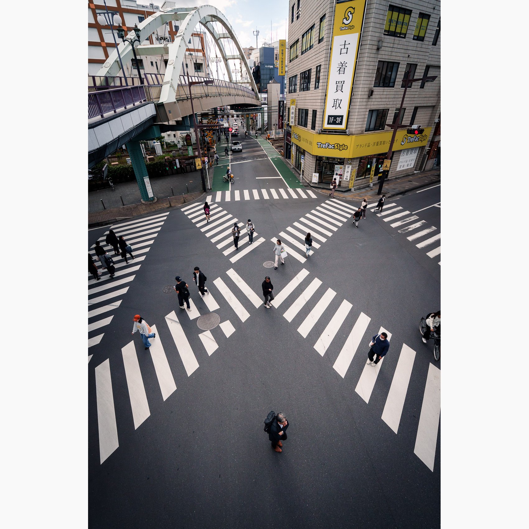 Overhead view of a Tokyo pedestrian crossing in Adachi, Japan, showing intersecting zebra crossings, commuters, and an elevated white bridge.