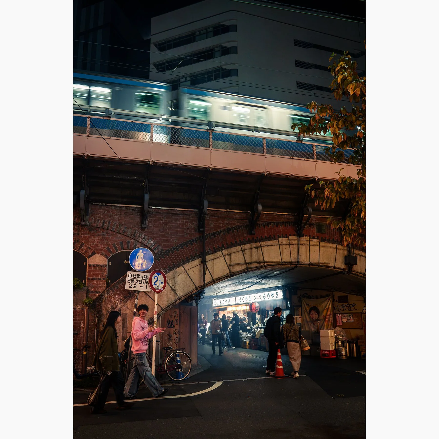 Tokyo night street scene with a train in motion above a glowing izakaya beneath the railway arches and pedestrians passing below.