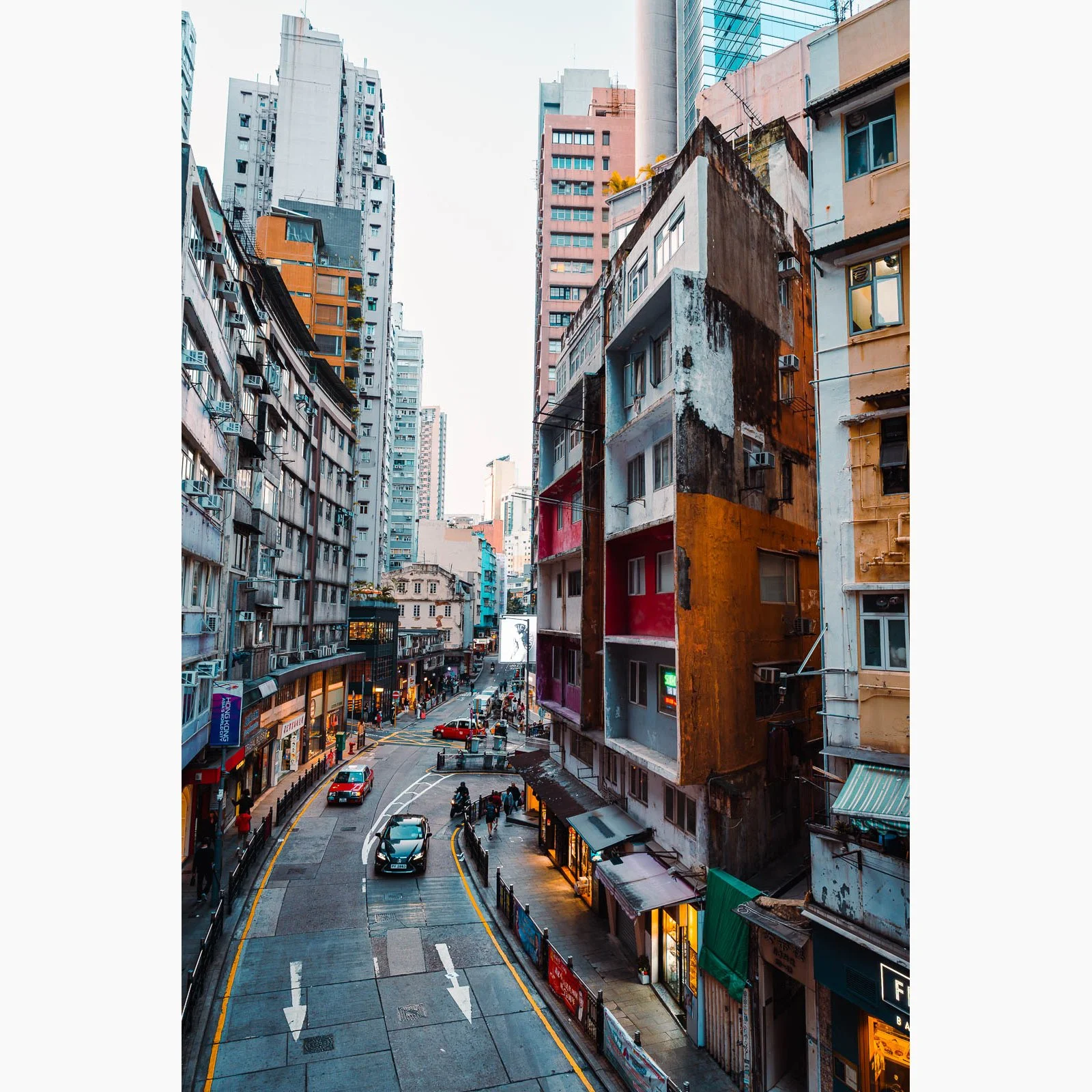Vertical Hong Kong street photography featuring dense apartment buildings, curved roadway, cars and warm city lights at dusk.