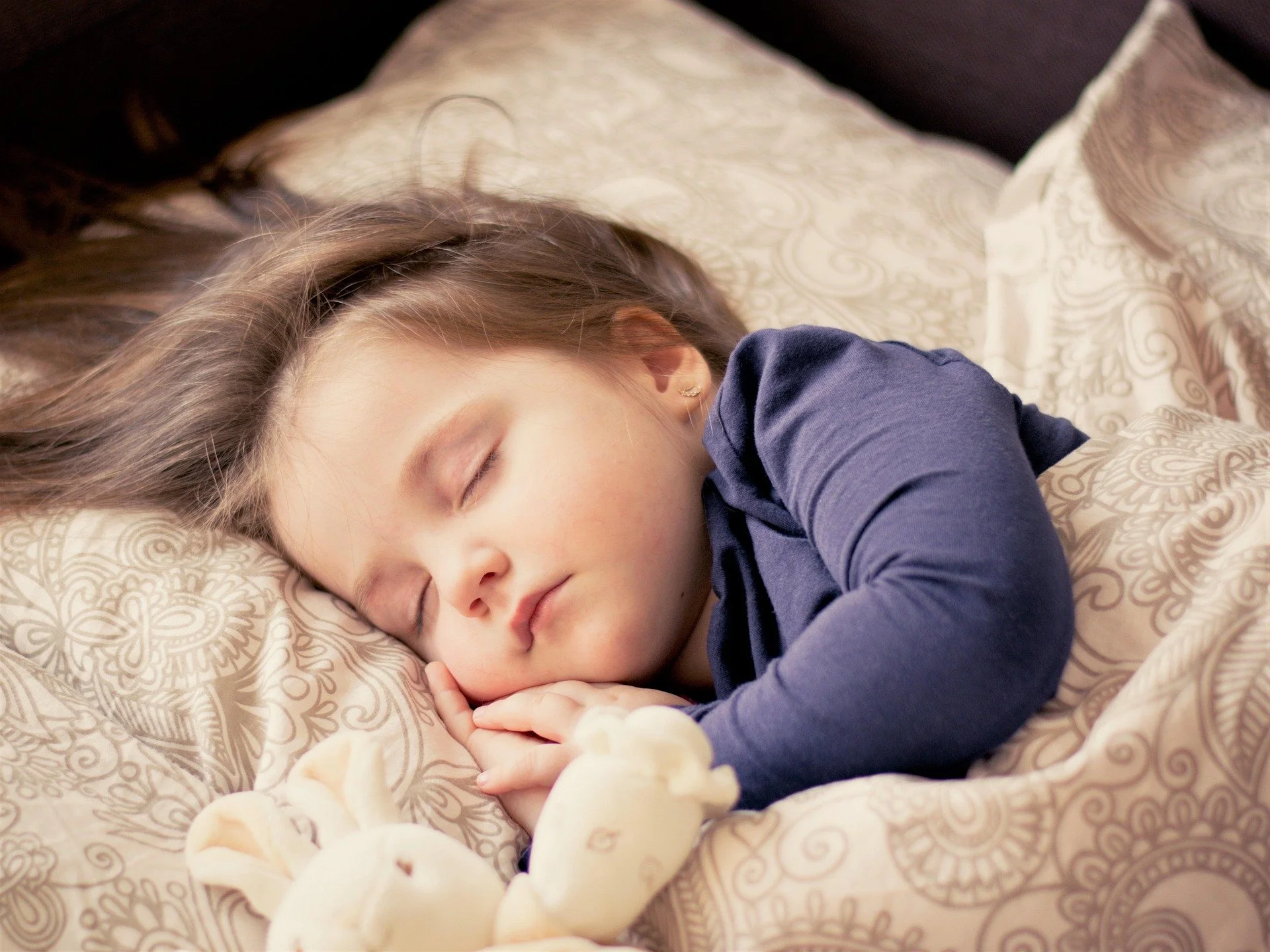 A young girl with long, light brown hair sleeping peacefully on a patterned bed with cream and beige bedding, wearing a dark blue long-sleeve shirt.
