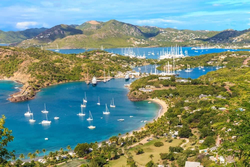 Scenic view of a marina with sailboats in a bay surrounded by green hills and distant mountains under a partly cloudy sky.