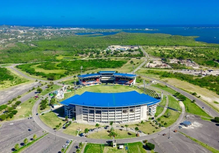 A large stadium with a blue roof located in a green area near water, with parking lots and roads surrounding it.