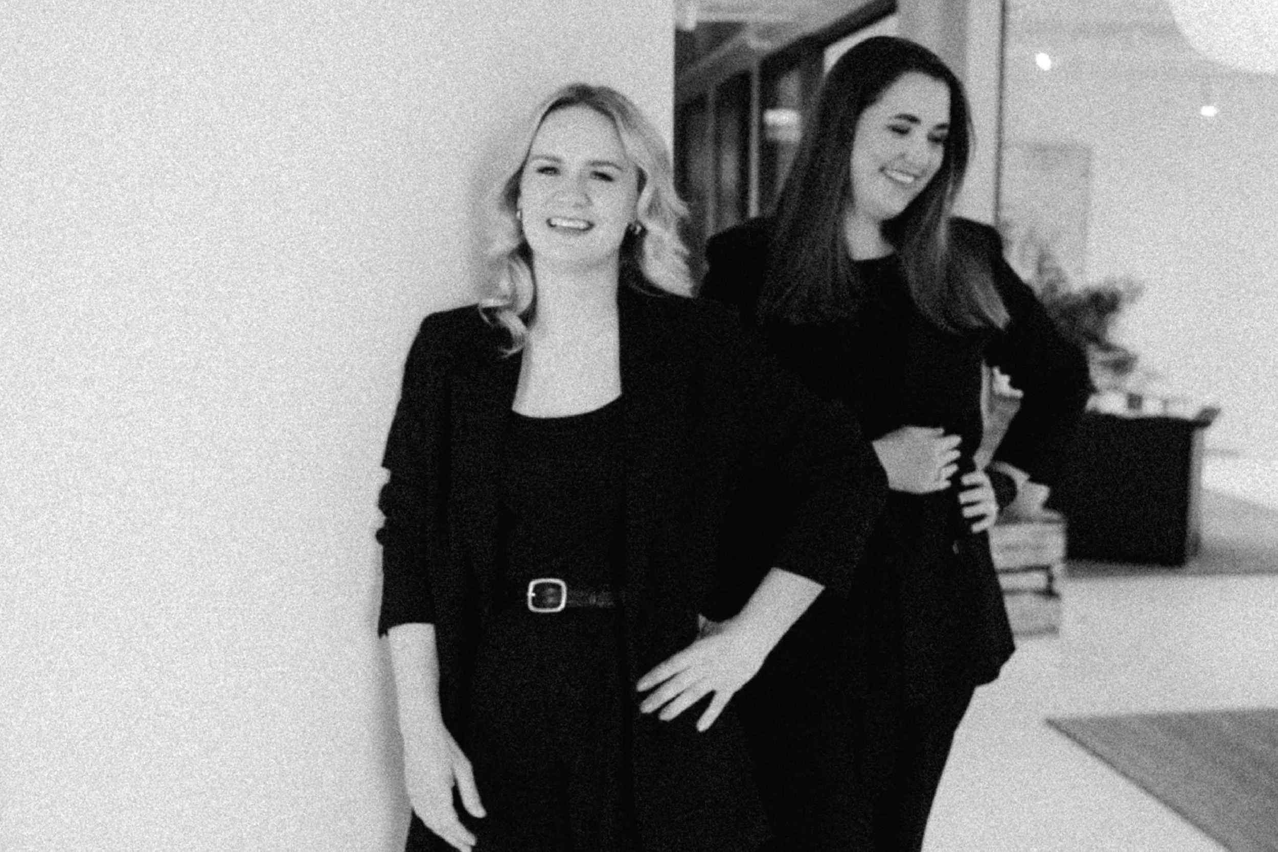 Two women standing indoors, laughing and smiling, dressed in black, with books and furniture in the background.