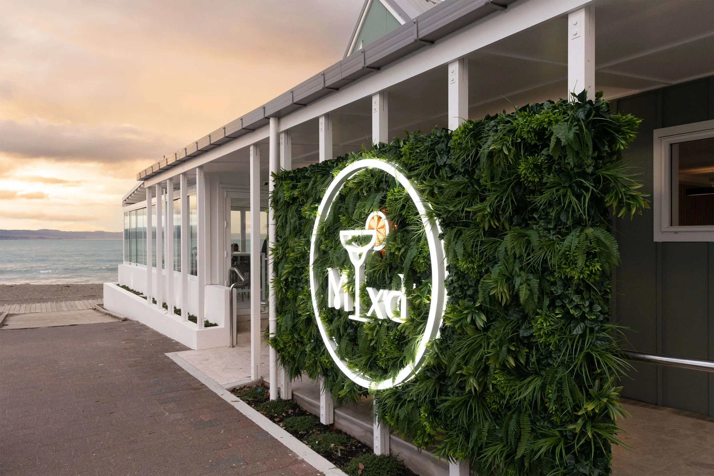 Exterior of a beachside restaurant or bar with a large neon sign reading "Dux" and a cocktail glass with an orange slice inside, surrounded by green plants on the wall. The scene is during sunset with the ocean and a cloudy sky in the background.