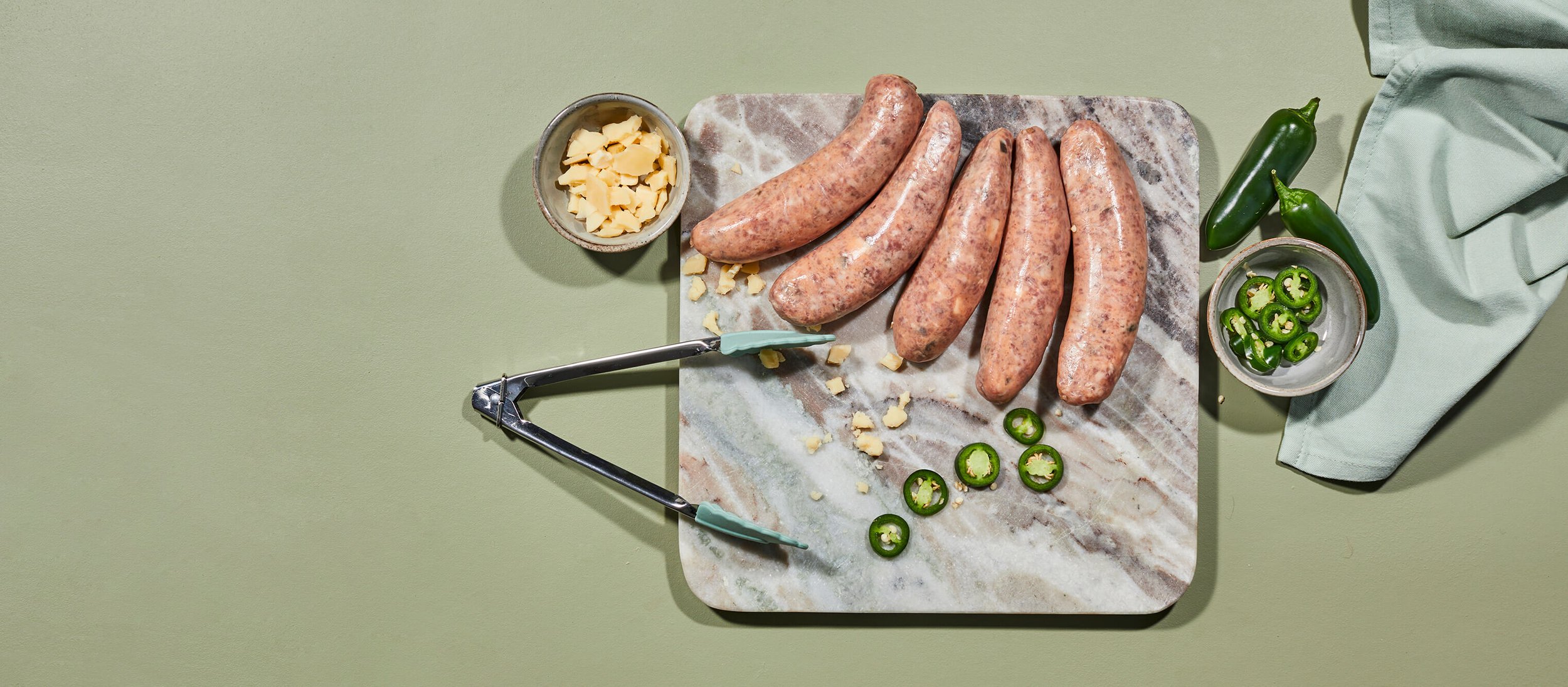 Raw sausages on a marble cutting board with sliced jalapeños, shredded cheese, and a pizza cutter, with fresh jalapeños and a green cloth on the side.