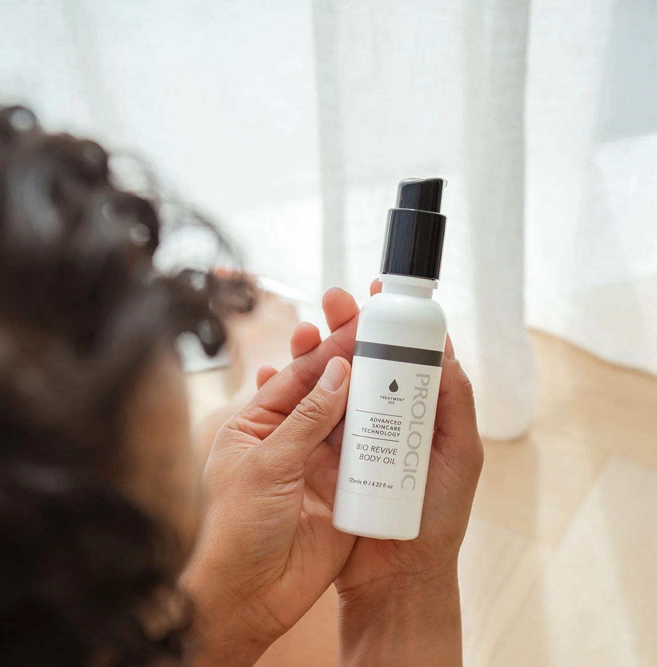 A person holding a white bottle of PROLOGIC Bio Revive Body Oil with a black pump dispenser, with curly hair visible in the foreground, indoors with sheer white curtains in the background.