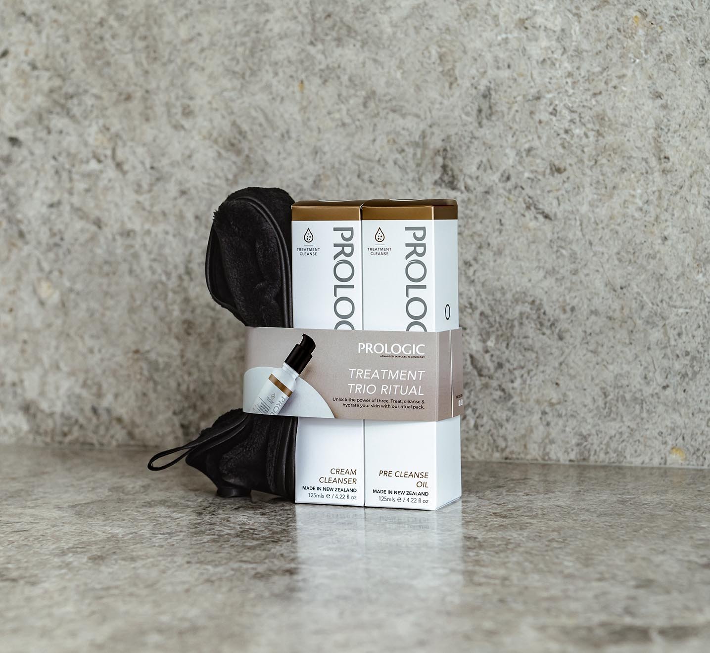 Prologic skincare set, including cream cleanser and pre cleanse oil, with a black facial brush in the background, on a gray textured surface against a gray textured wall.