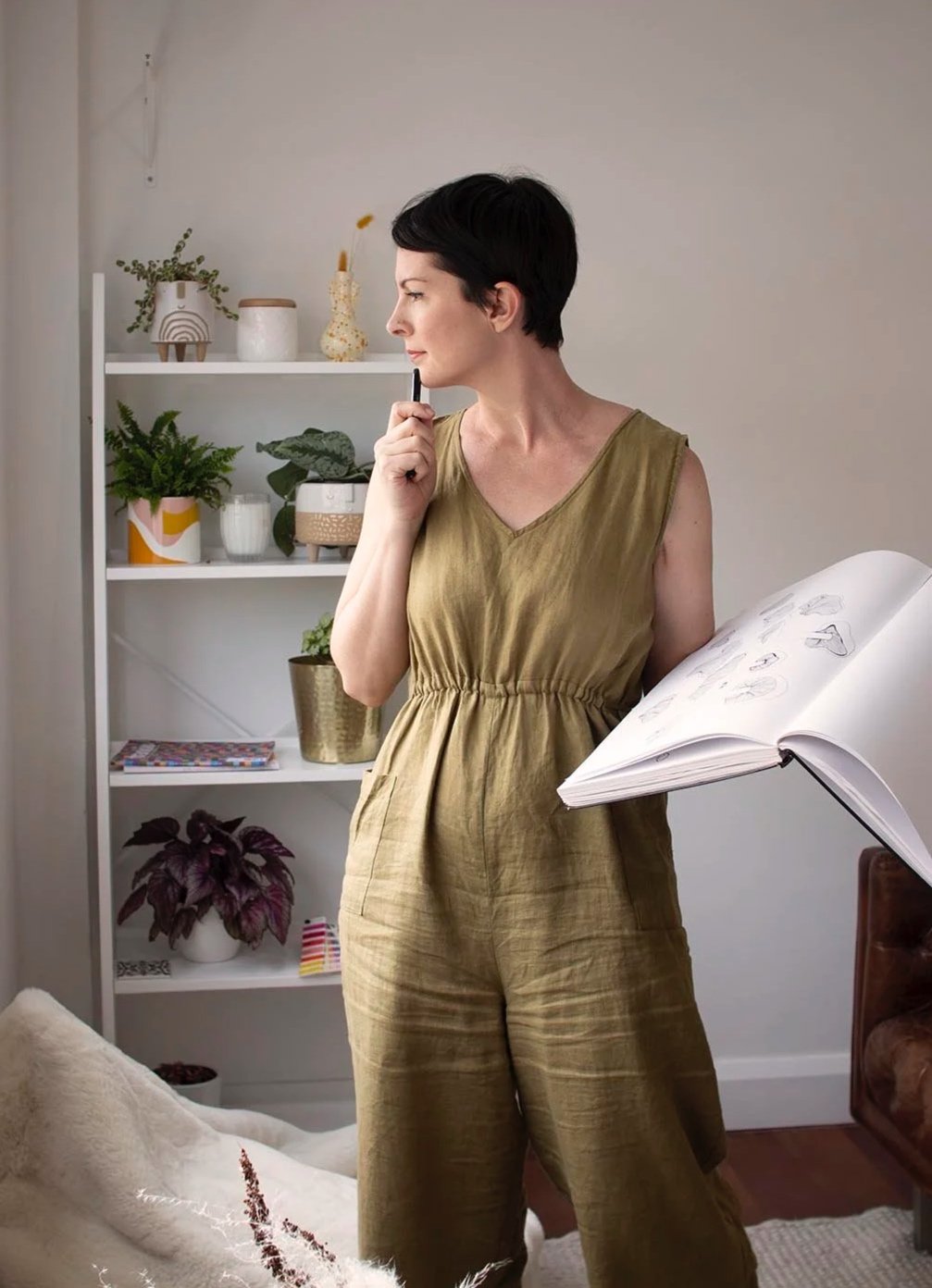 A woman with short dark hair wearing a gold-colored sleeveless jumpsuit stands indoors, holding a black marker near her chin while looking thoughtfully to her left. She has a sketchbook open in her left hand. Behind her is a white bookshelf with assorted potted plants and decorative items, and a beige couch with throw pillows is partially visible in the foreground.
