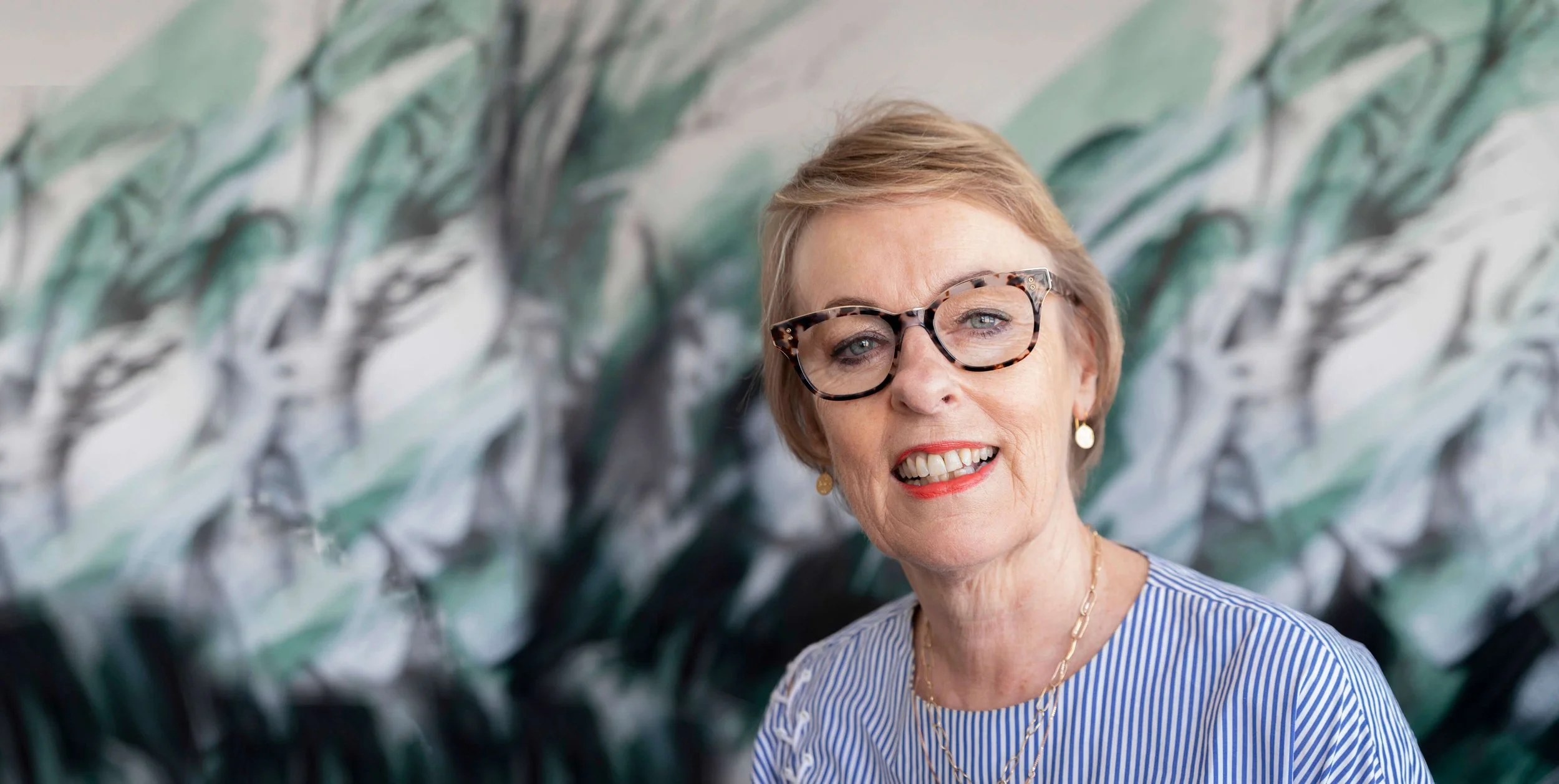 An elderly woman with short blonde hair, wearing tortoise shell glasses, red lipstick, a blue and white striped blouse, gold earrings, and layered gold necklaces, smiling in front of a colorful abstract background.