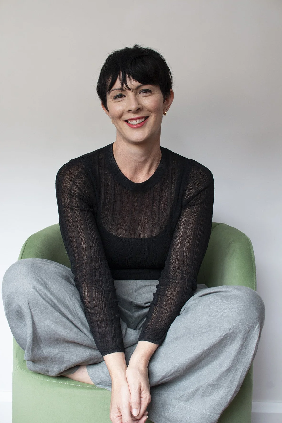 A woman with short black hair, smiling, wearing a black sheer long-sleeve top and gray pants, sitting cross-legged on a green chair against a plain white wall.