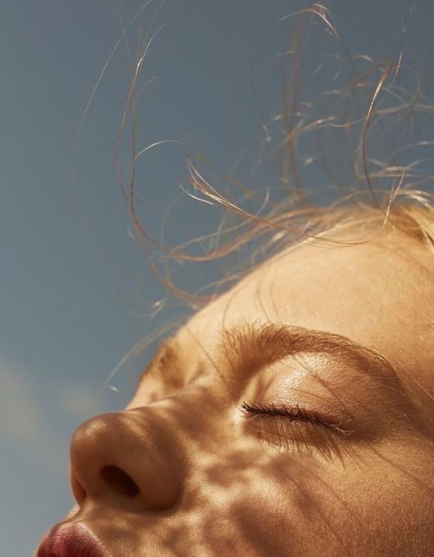 Close-up of a woman's face with closed eyes and wind-blown hair against a blue sky.