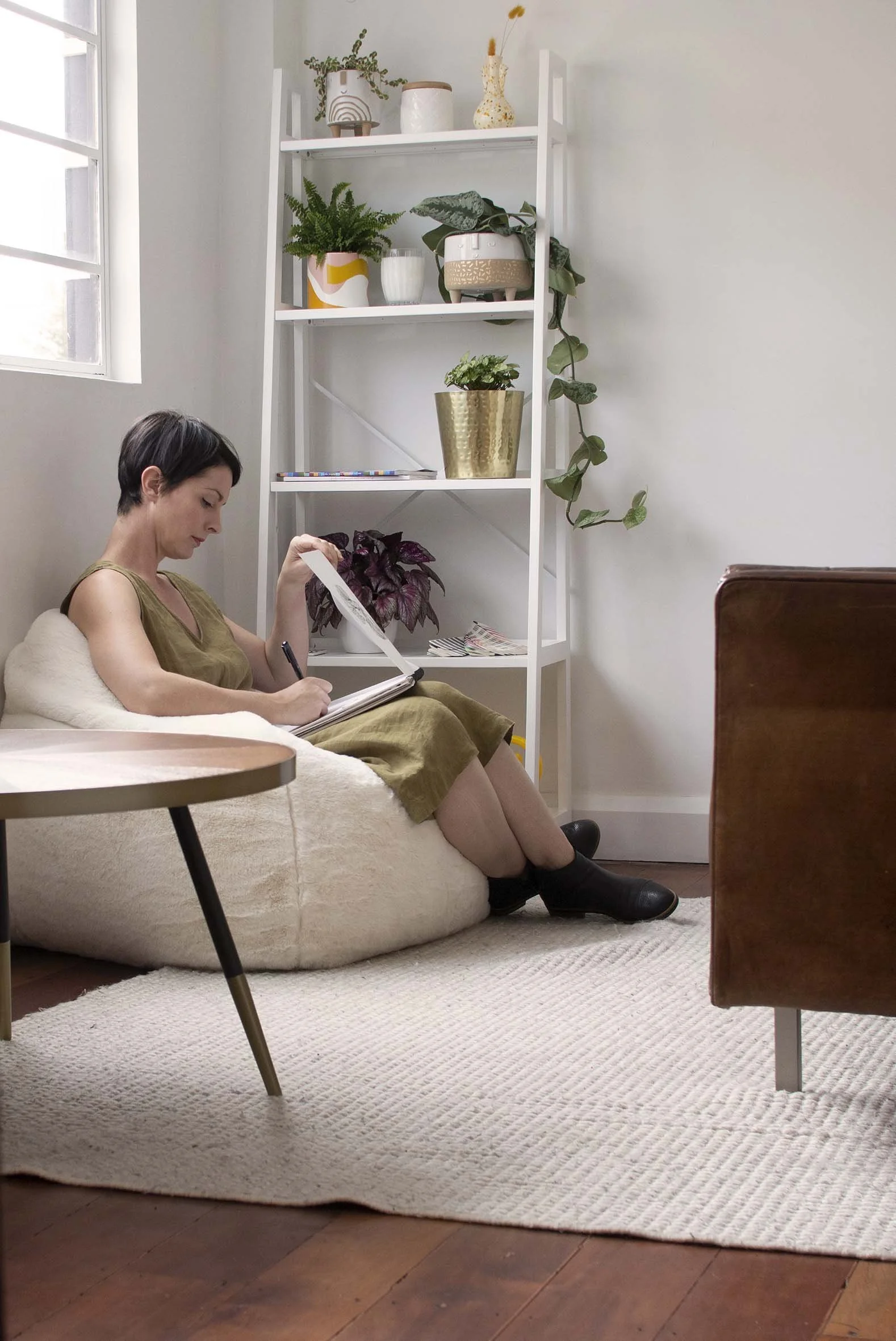 Woman sitting on a bean bag chair, taking notes with a pen and notebook in a cozy living room with a white bookshelf behind her, decorated with plants and vases.