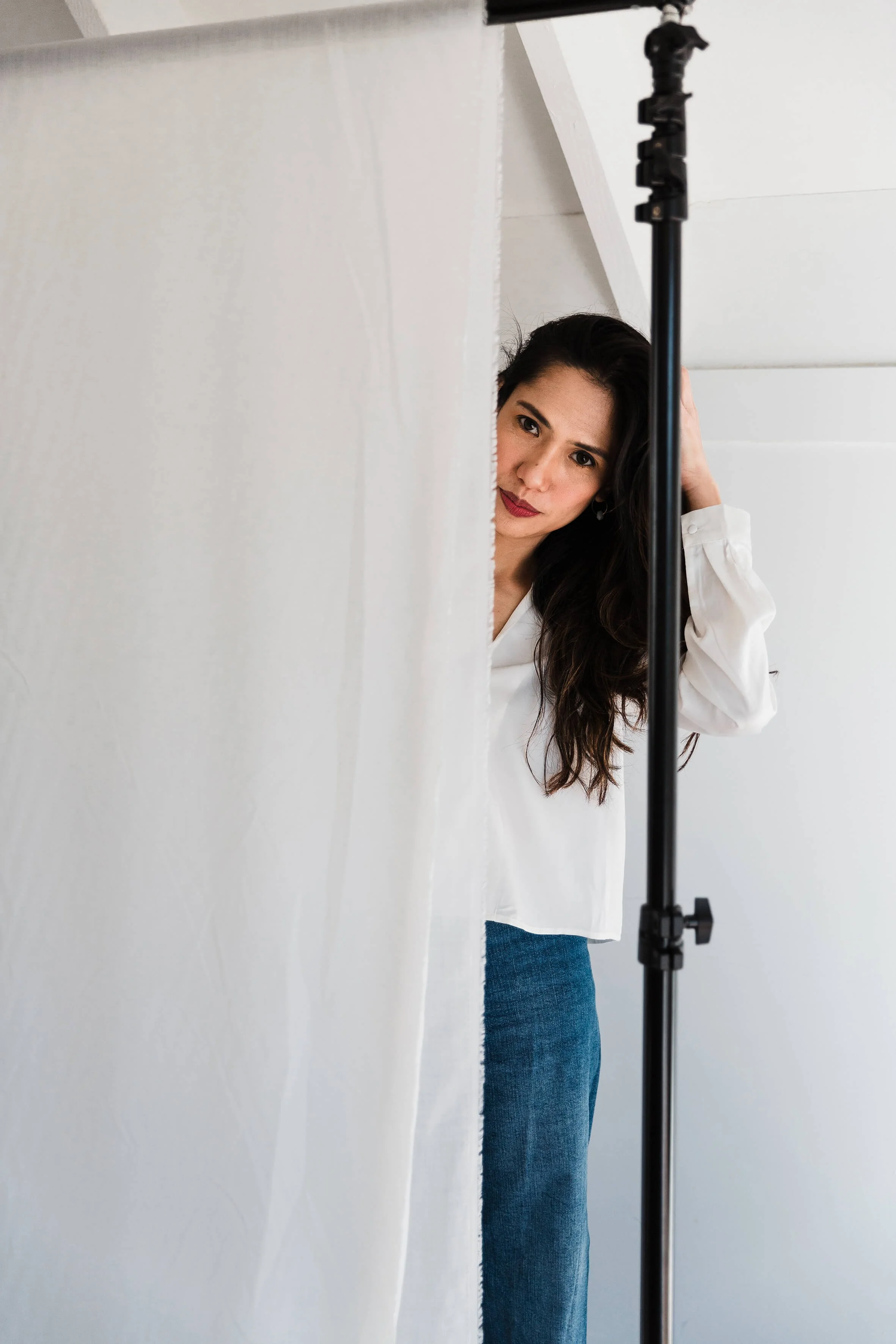 A woman with long dark hair wearing a white blouse and blue jeans is peeking from behind a white fabric backdrop while holding her hair with one hand. There is a black tripod stand in front of her.