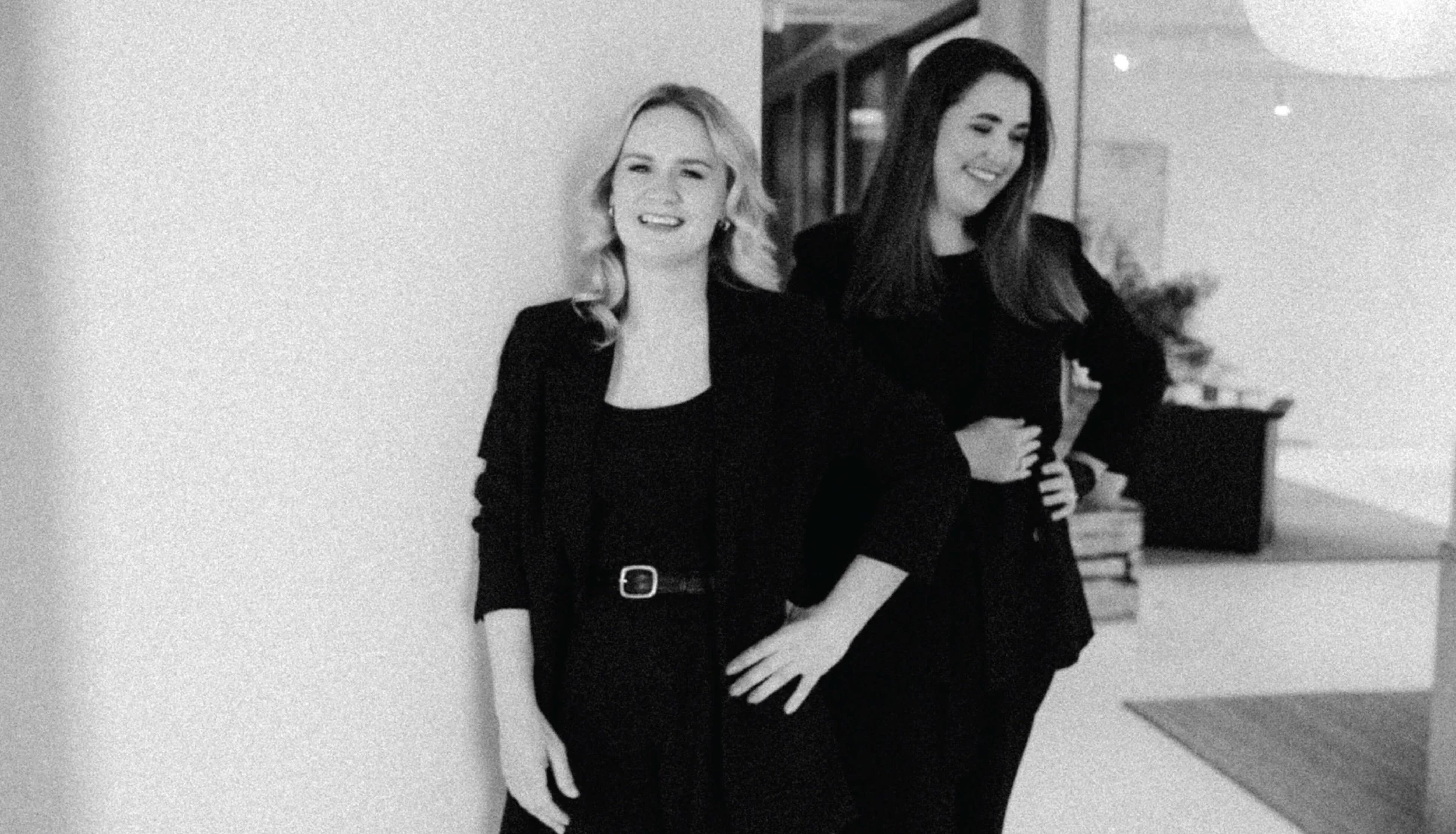 Two women standing indoors, smiling and posing for the photo, with a plain wall on the left and a room with furniture and plants in the background.