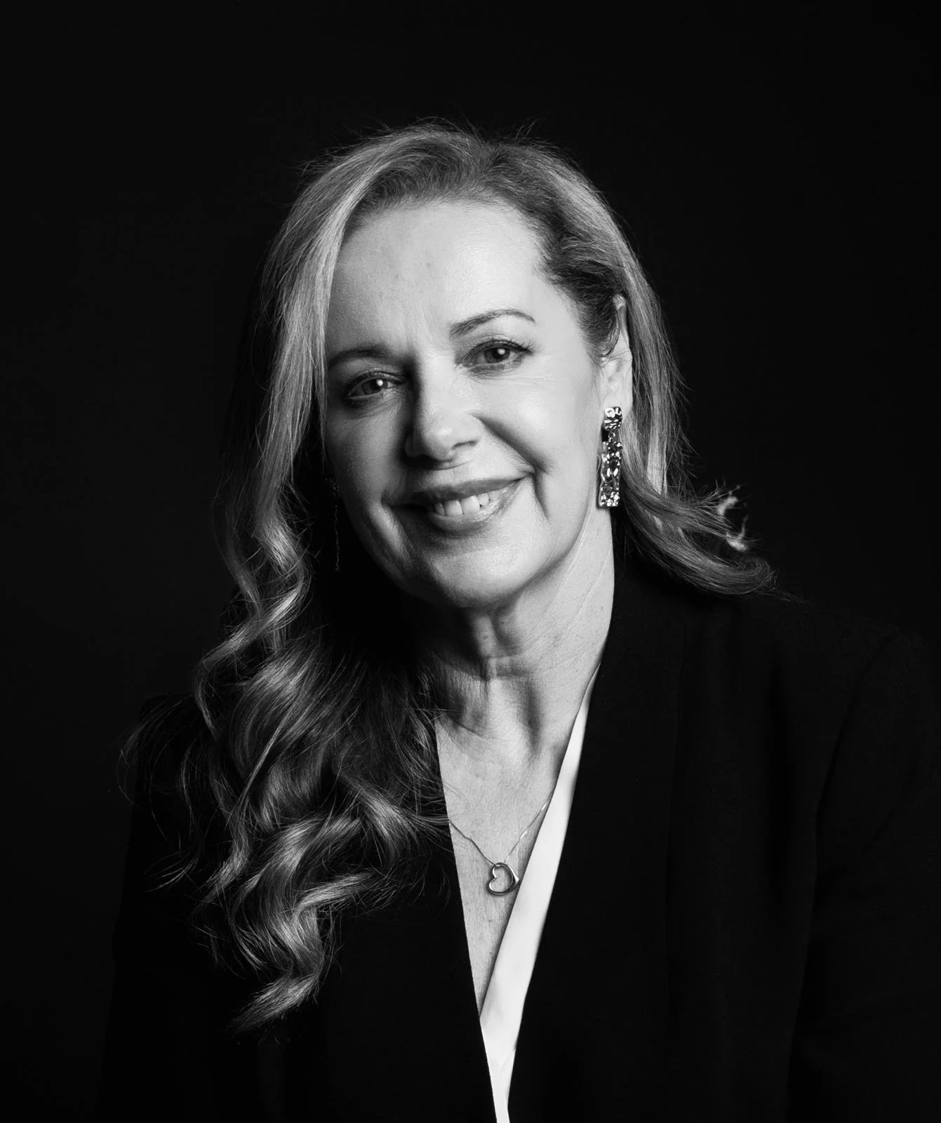 Black and white portrait of a smiling middle-aged woman with long, wavy hair, wearing earrings and a necklace, against a dark background.