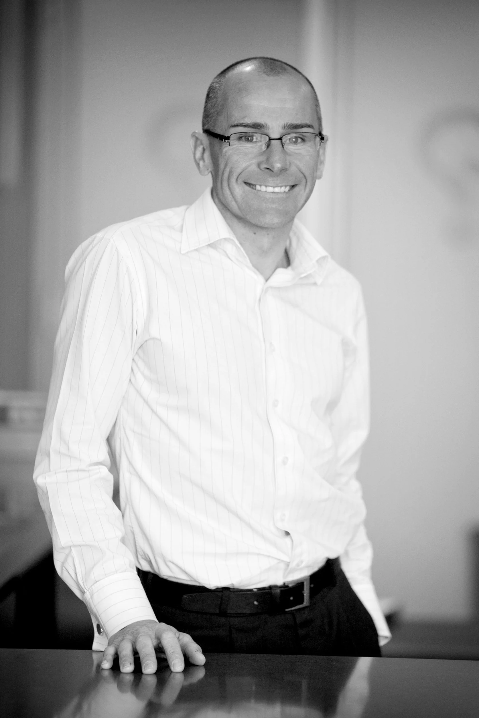 A man wearing glasses and a striped white shirt, smiling, standing with his hand resting on a table in an office or professional setting.