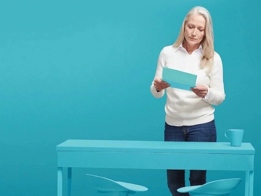 A woman with long gray hair, wearing a white sweater and dark jeans, standing behind a light blue table with a matching mug, reading from a piece of paper against a solid blue background.