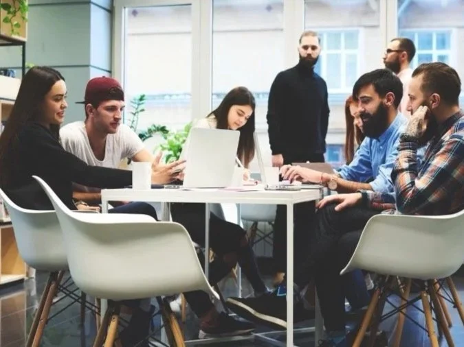 Group of seven young adults in a modern office, some sitting around a table with laptops, two standing near the window, engaging in a discussion.