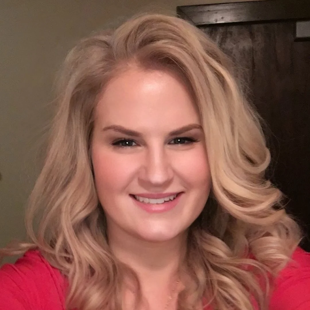 A smiling young woman with long, wavy blonde hair, wearing a red top, standing indoors with a dark wooden door in the background.