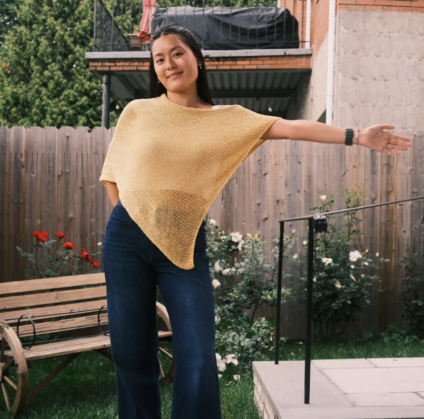A woman standing in a backyard with a wooden fence, flowers, and a brick building in the background. She is wearing a yellow knit top and blue jeans, with her right arm extended outward.
