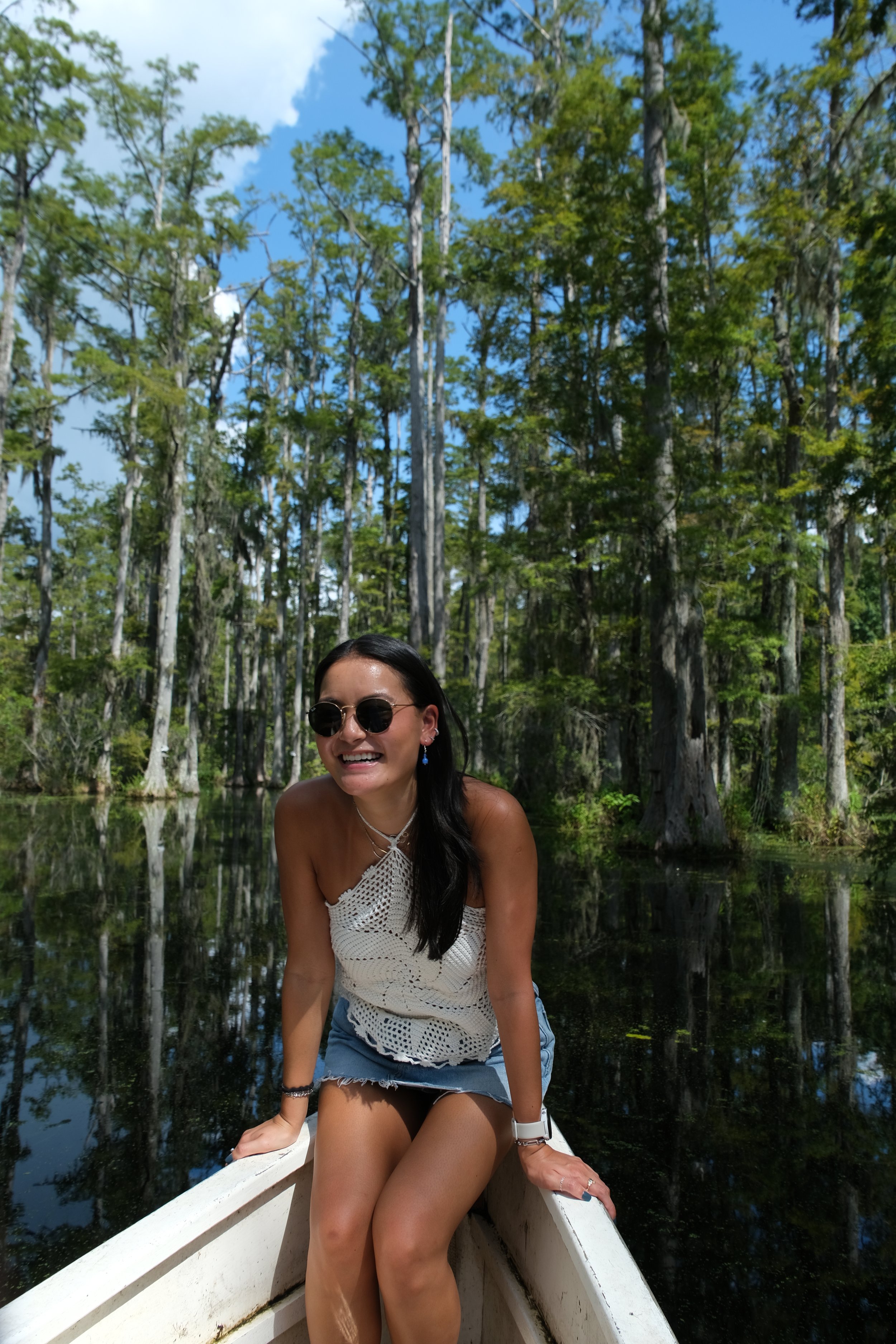 A woman with long dark hair wearing sunglasses, a white crocheted top, and denim shorts, sitting in a boat on a calm body of water surrounded by tall trees and green foliage under a blue sky.