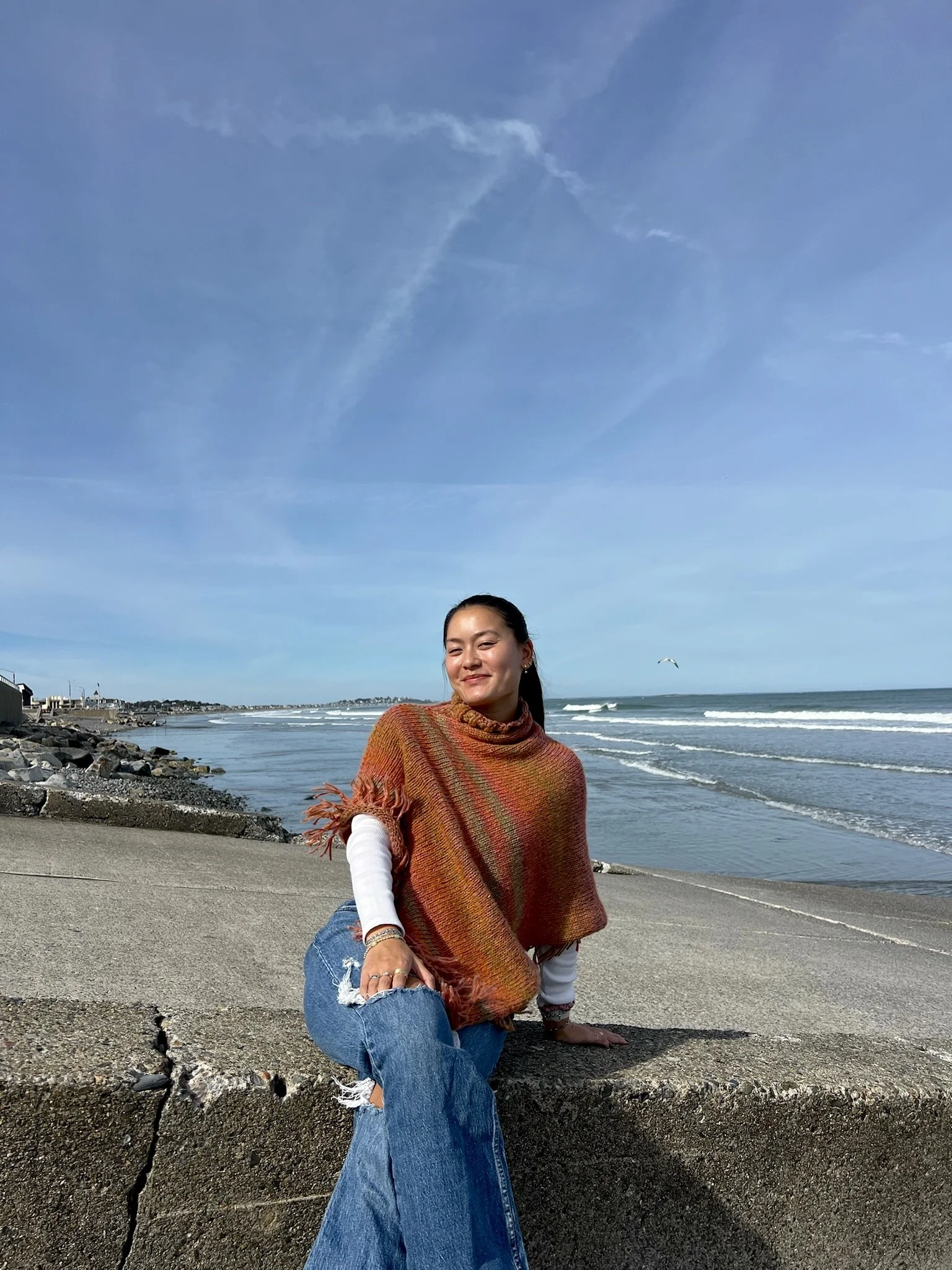 woman sitting on a concrete ledge at the beach, wearing a colorful poncho and ripped jeans, with the ocean and blue sky in the background.