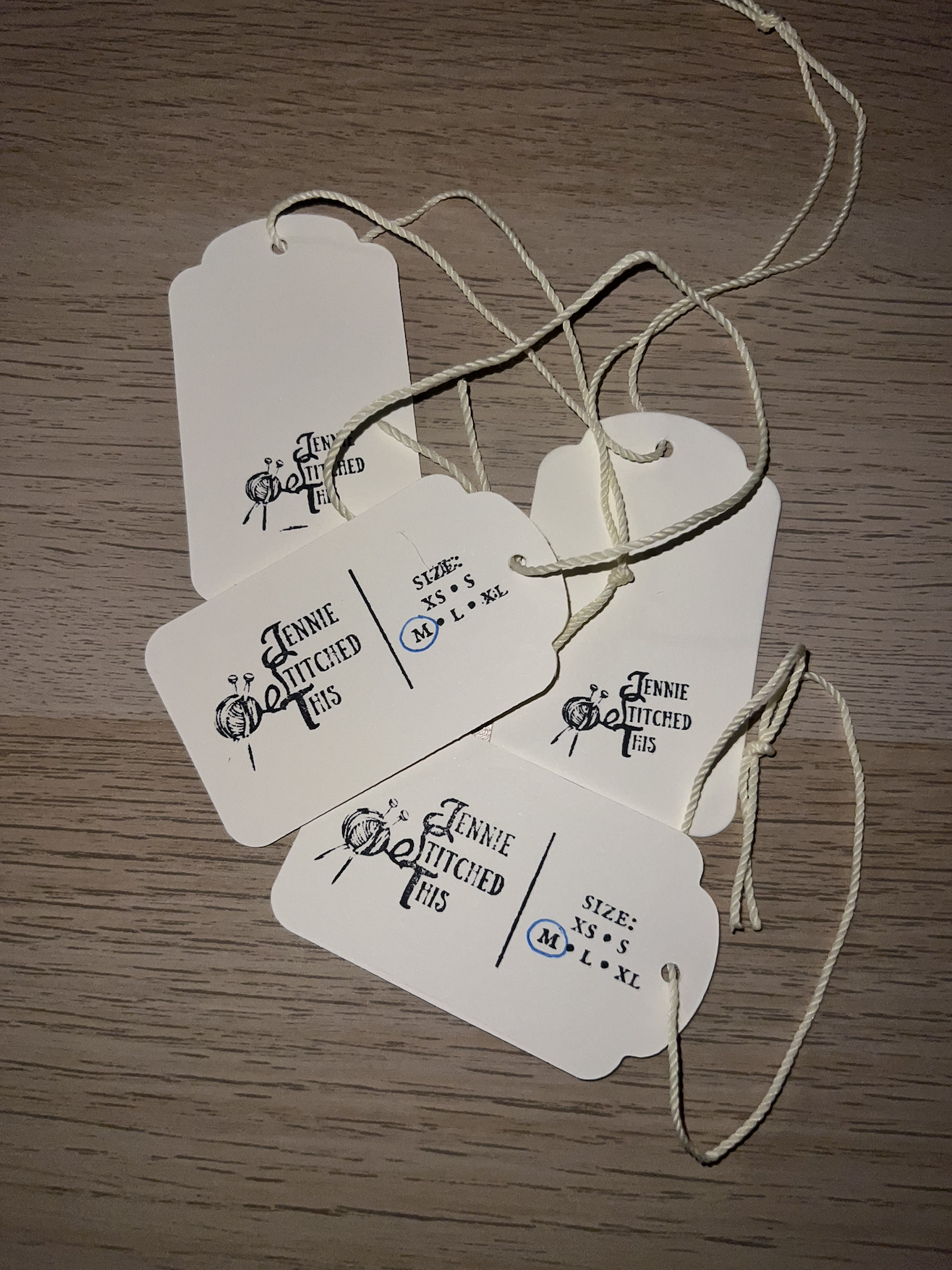 A bundle of tags with the label 'Jennie Titched Tais' and size options XS, S, M, L, XL. The tags are attached to a white string and placed on a wooden surface.