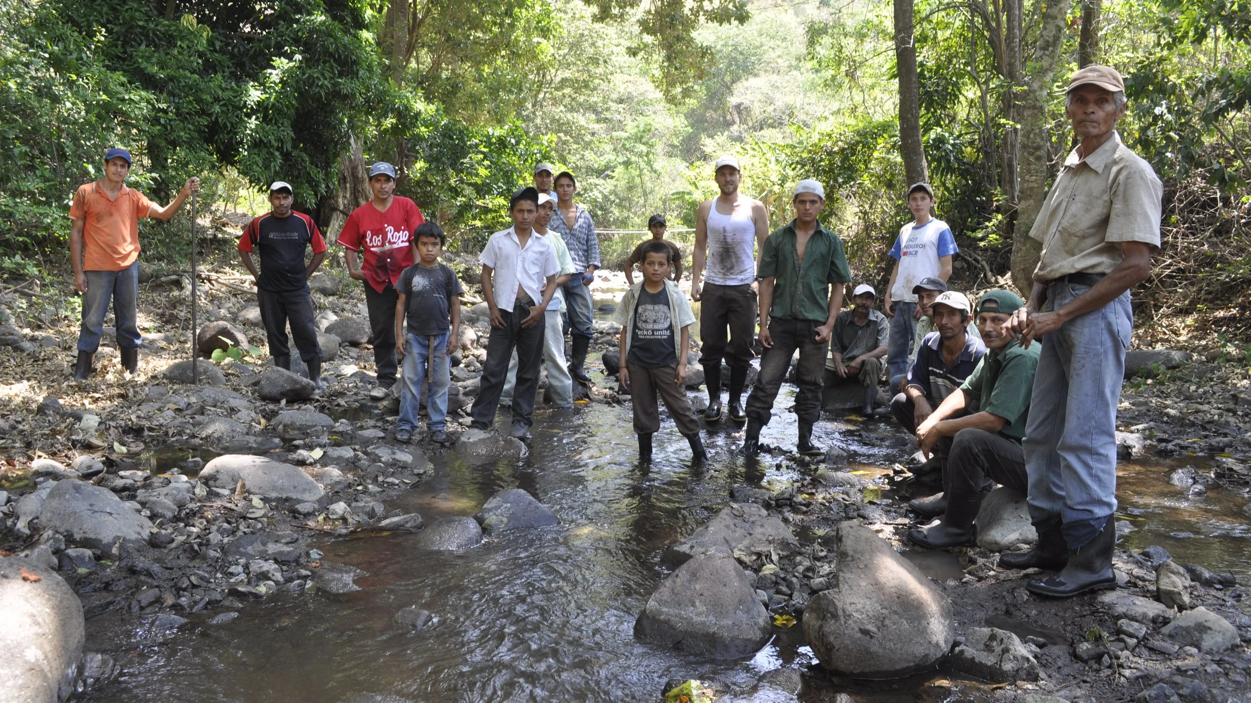 A group of people, including children and adults, are standing in a shallow rocky stream in a dense forest. Some are wearing boots and casual outdoor clothing, and they appear to be exploring or working in the natural environment.