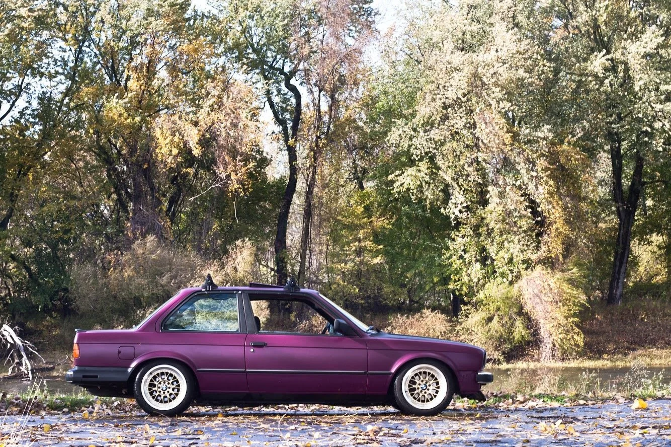 A purple vintage car parked on a cobblestone street beside a river, with trees showing autumn foliage in the background.