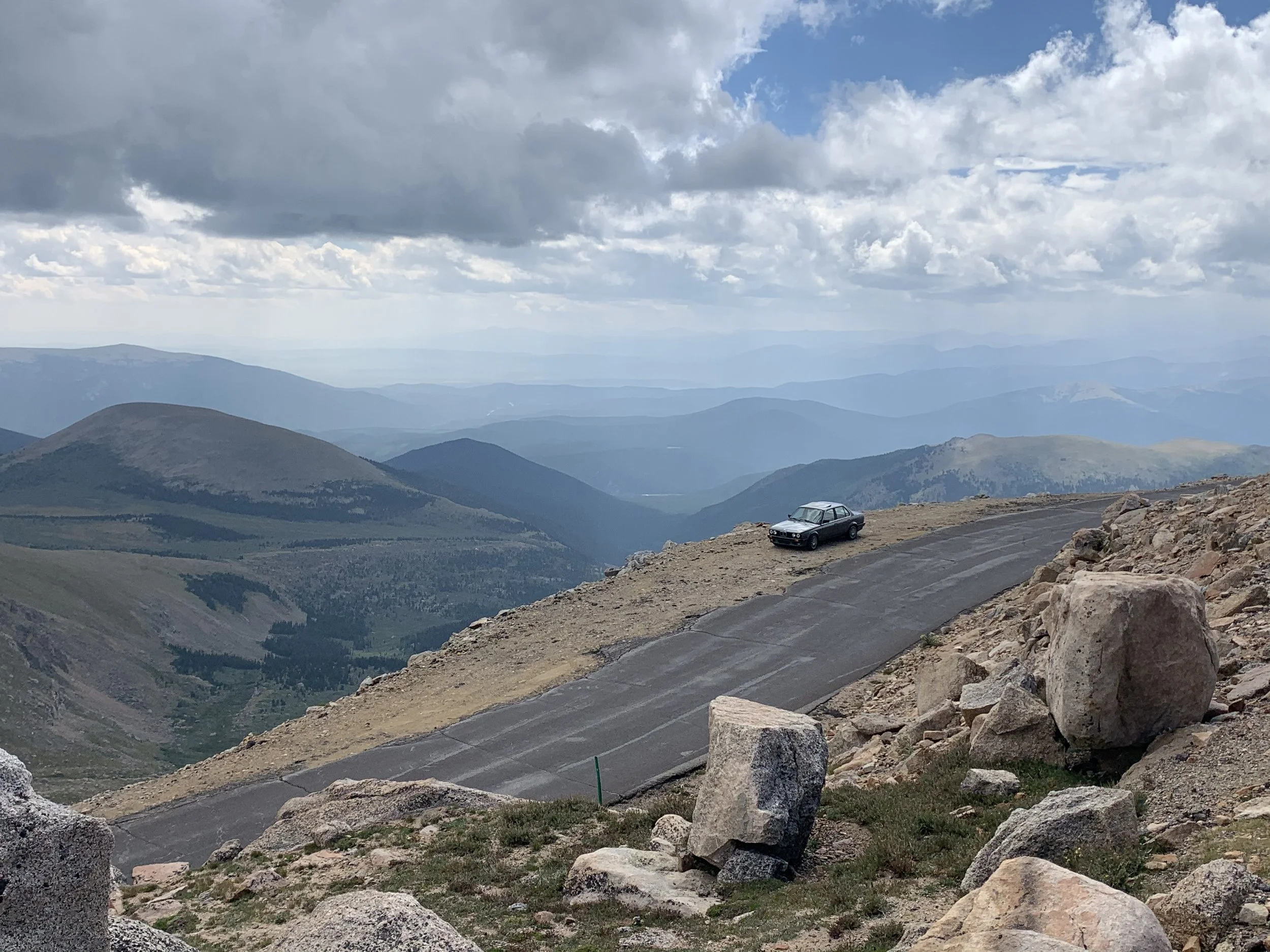 A black car parked on the side of a mountain road with a vast view of mountain ranges and a cloudy sky in the background.