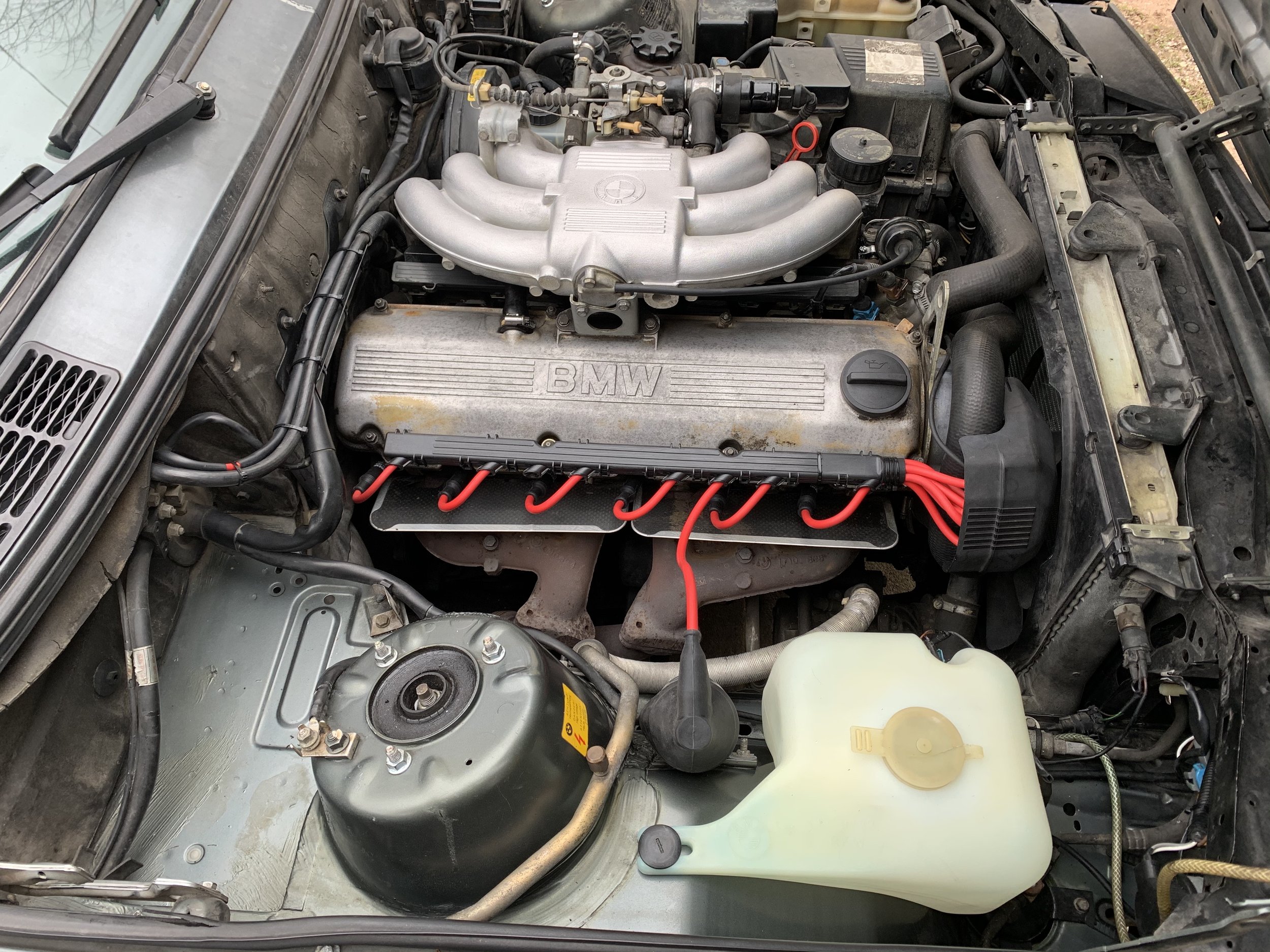 Inside view of a car's engine bay showing a BMW engine with red ignition wires, various hoses, and fluid reservoirs.