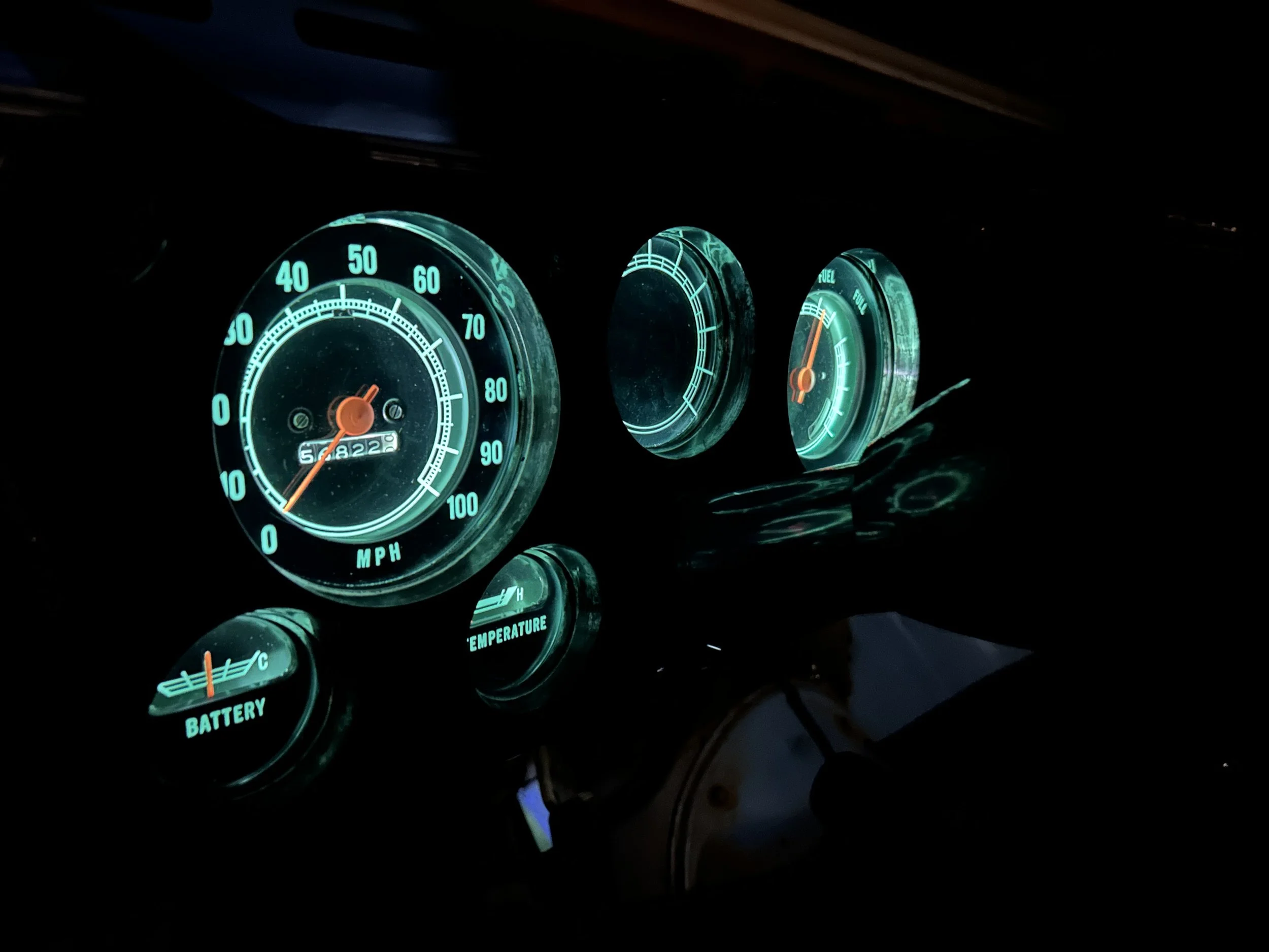 Dashboard of a vehicle with illuminated gauges, including speedometer, fuel gauge, battery, and temperature gauge, all glowing with a teal light in the dark.