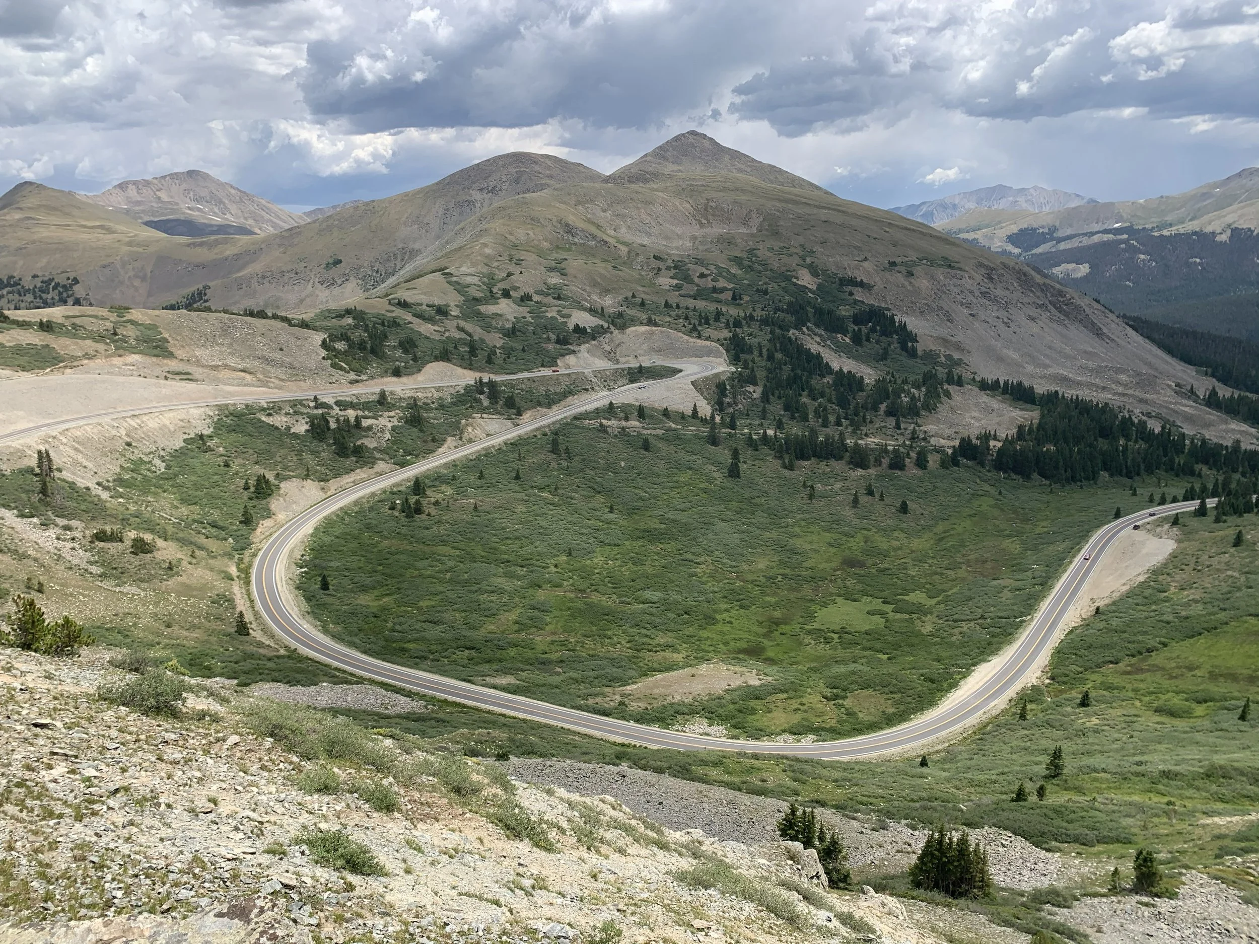 Winding mountain road through green hills and rocky terrain, with mountains and cloudy sky in the background.