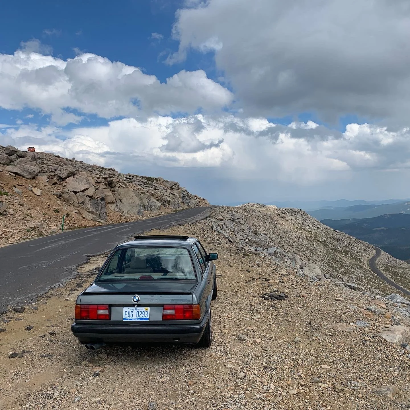 A black BMW parked on a gravel shoulder overlooking a mountain landscape with a winding road and cloudy sky.