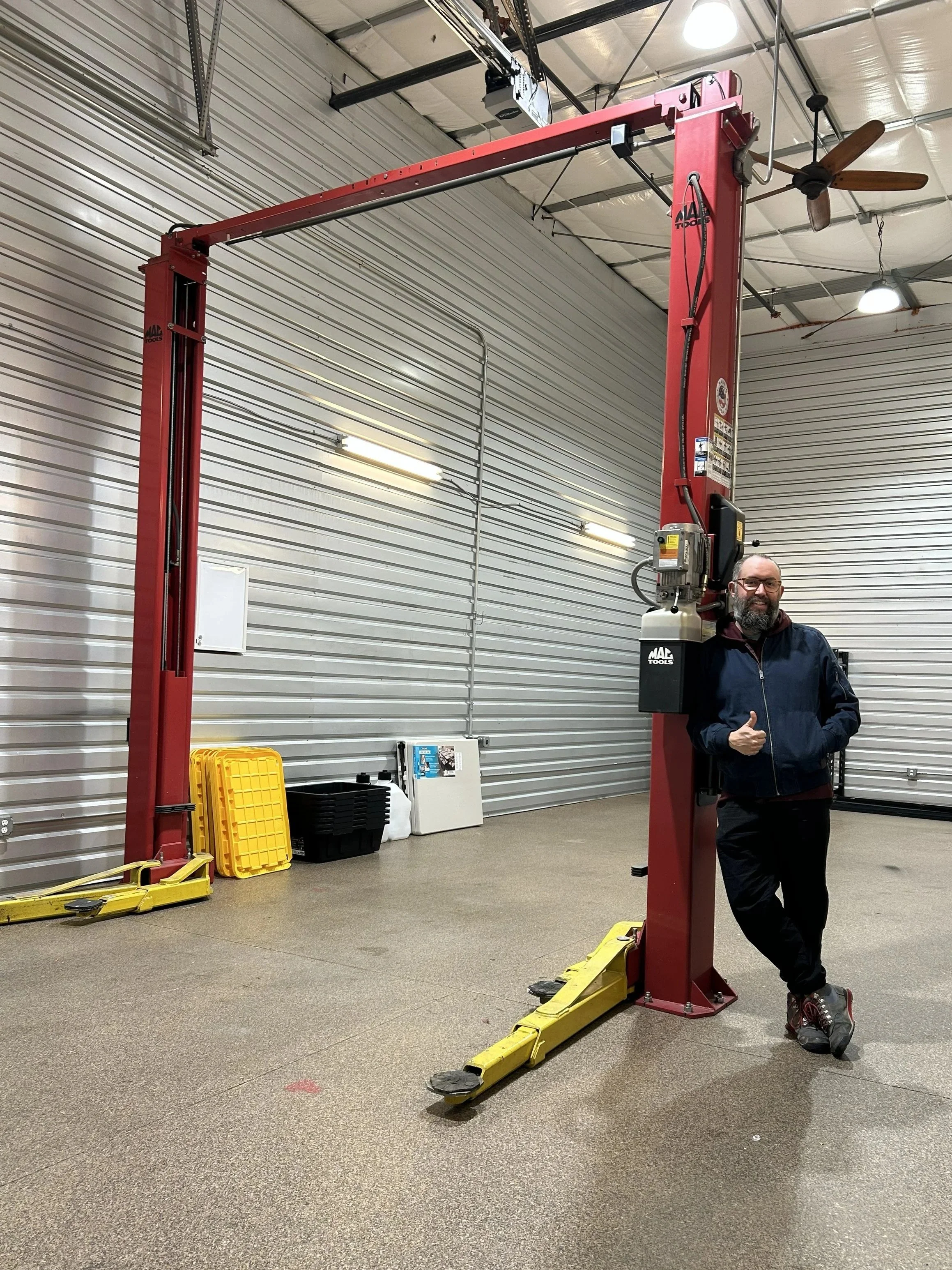 A man standing next to a large red automotive lift inside a garage or workshop, giving a thumbs-up.
