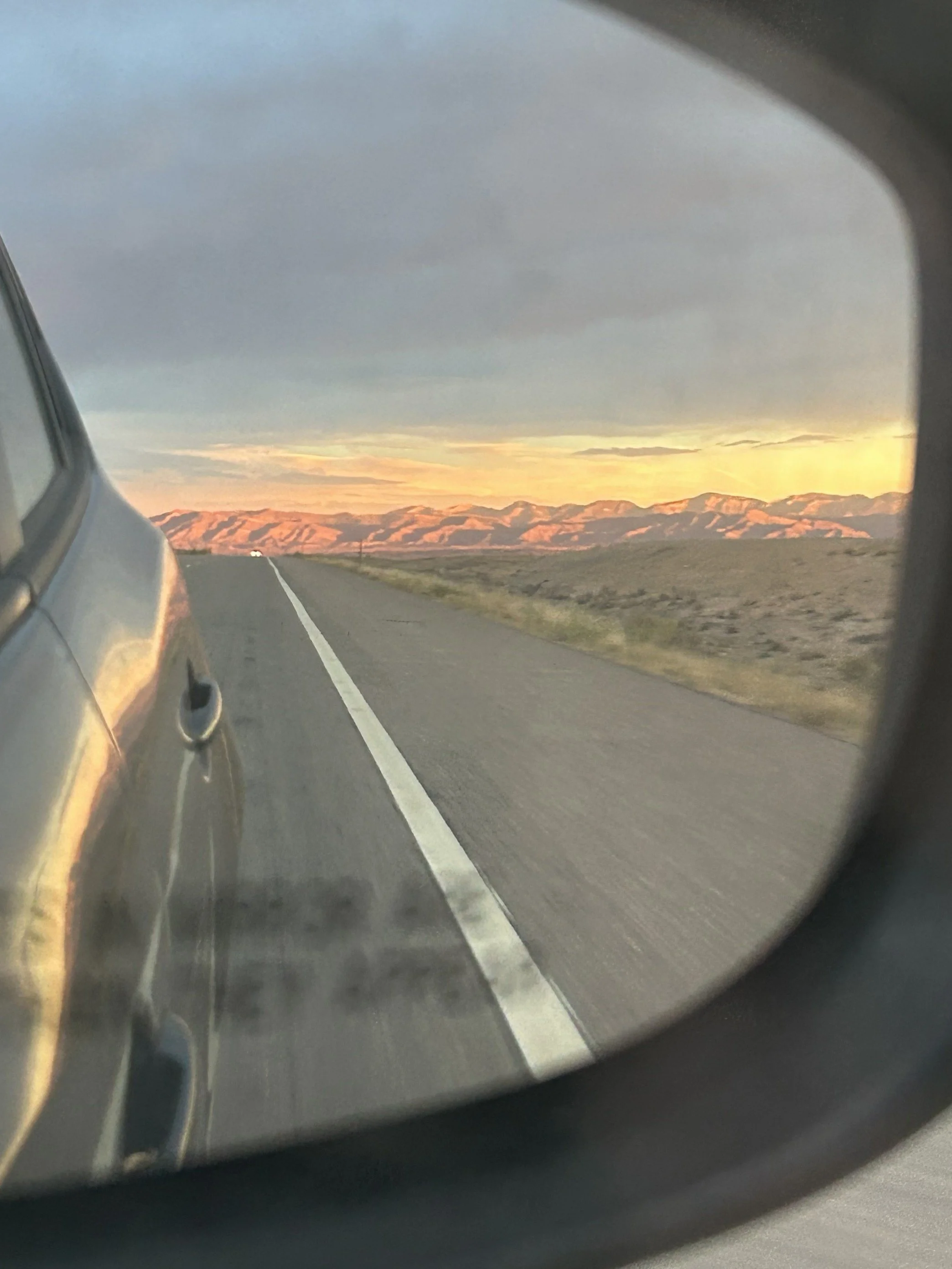 View of a road and distant mountains captured through a car's side mirror during sunset.