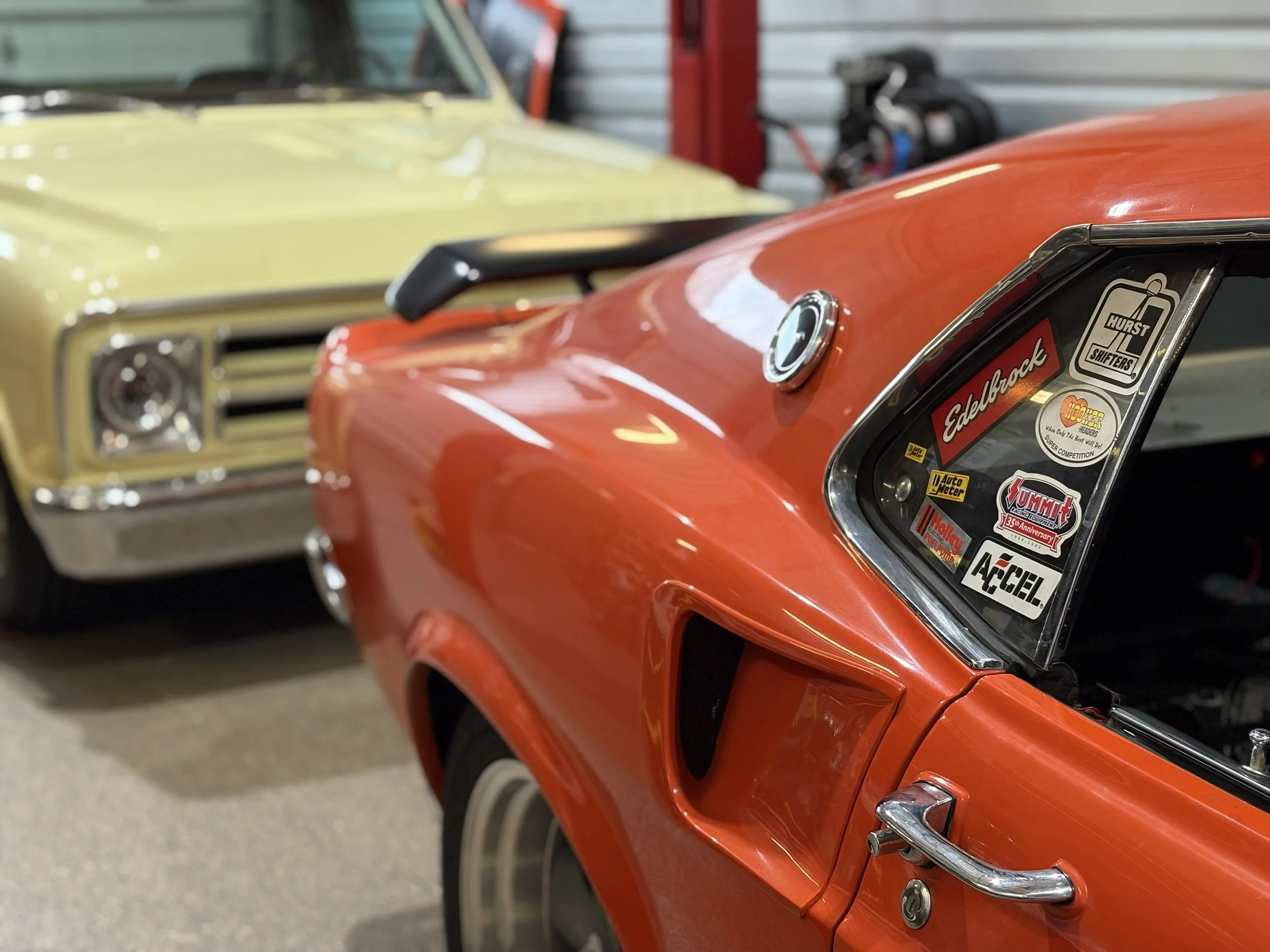 Close-up of an orange vintage race car with stickers on the window, behind a yellow classic car in a garage or showroom.