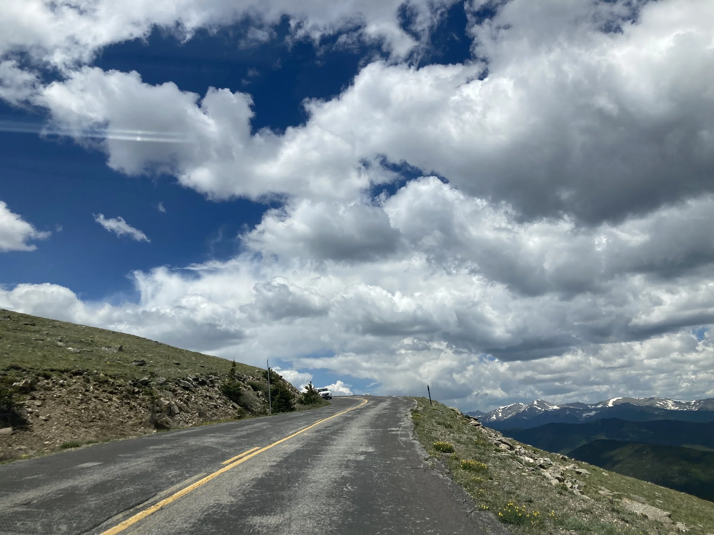 Rugged mountain road curving uphill through green, rocky landscape with snow-capped peaks in the distance under a partly cloudy sky.