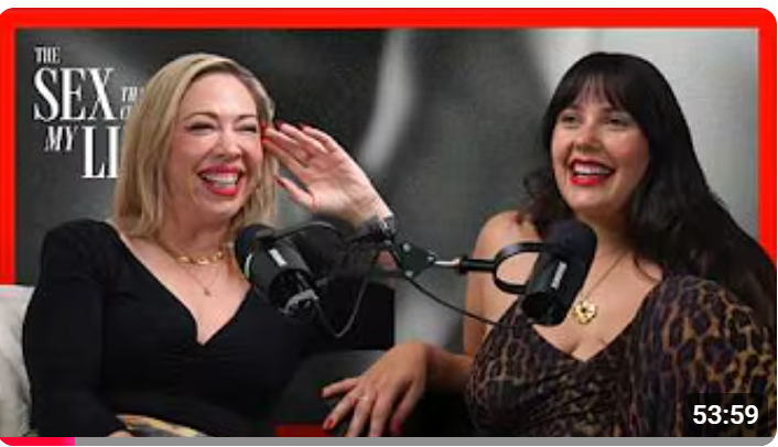 Two women sitting and laughing during a podcast recording with microphones, with a poster in the background that reads "The Sex that changed My Life."
