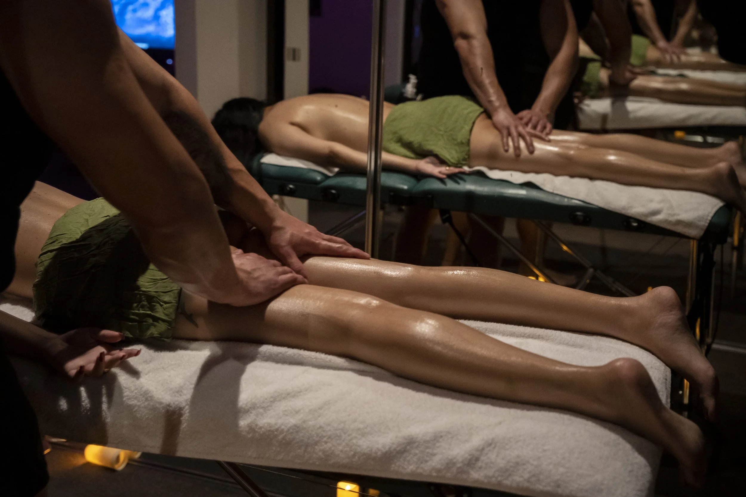 A person receiving a massage while lying face down on a massage table, with several others also on massage tables in a dimly lit massage room.