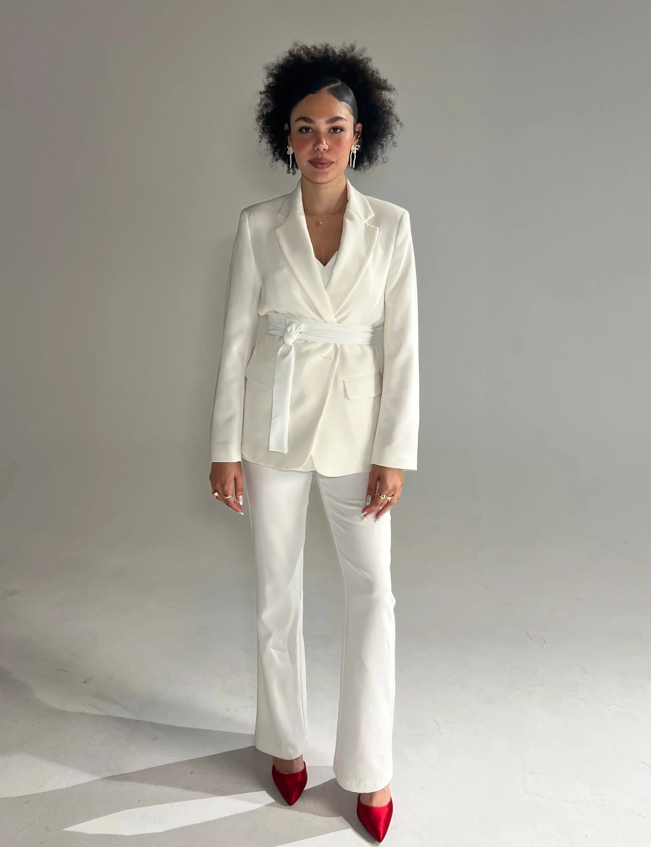 A woman with curly hair wearing a white pantsuit, red high heels, and gold jewelry, standing against a plain light-colored wall.