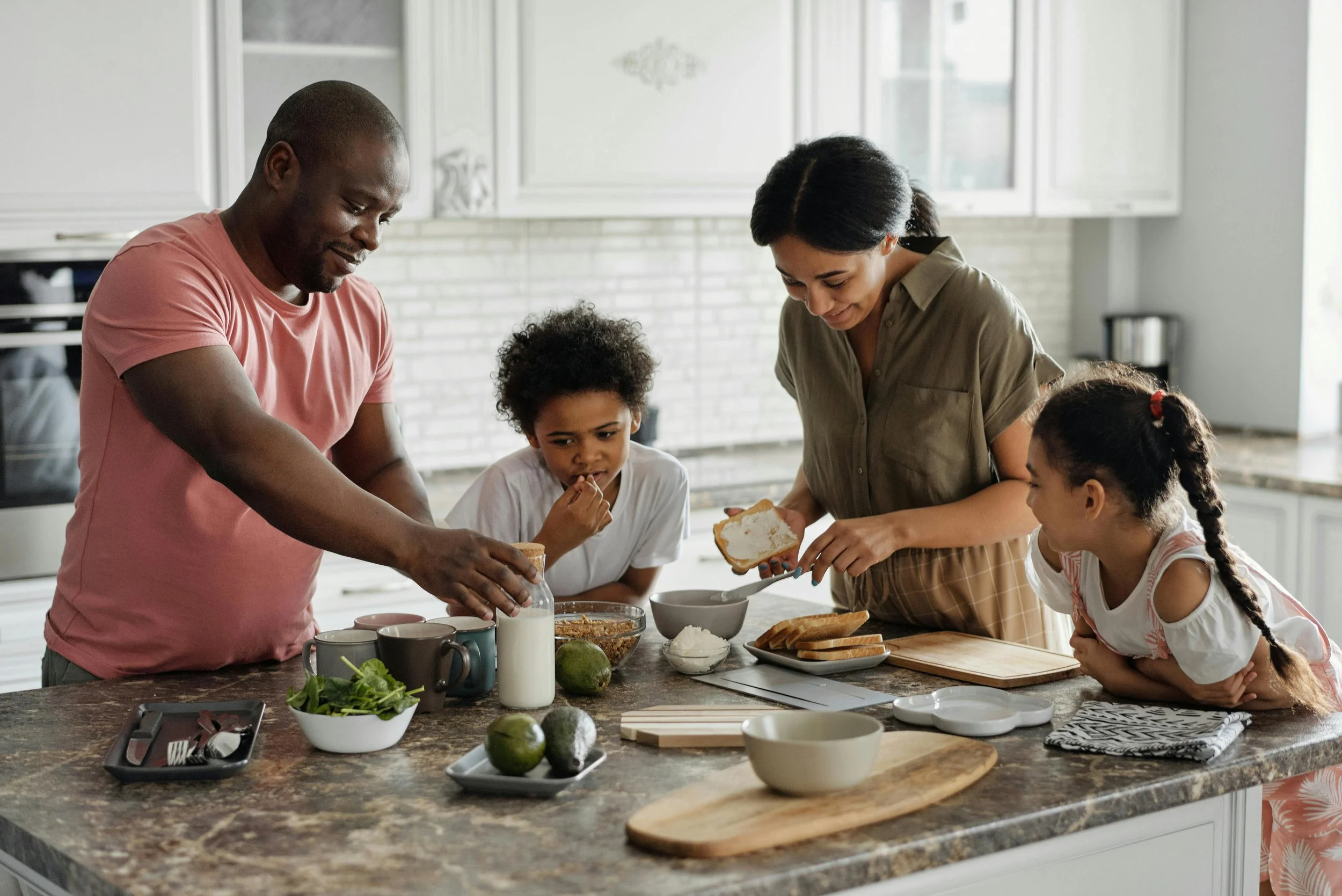 Family of five preparing breakfast in a kitchen, including a man pouring milk, a woman spreading butter on bread, two children watching, and various breakfast items on the counter.