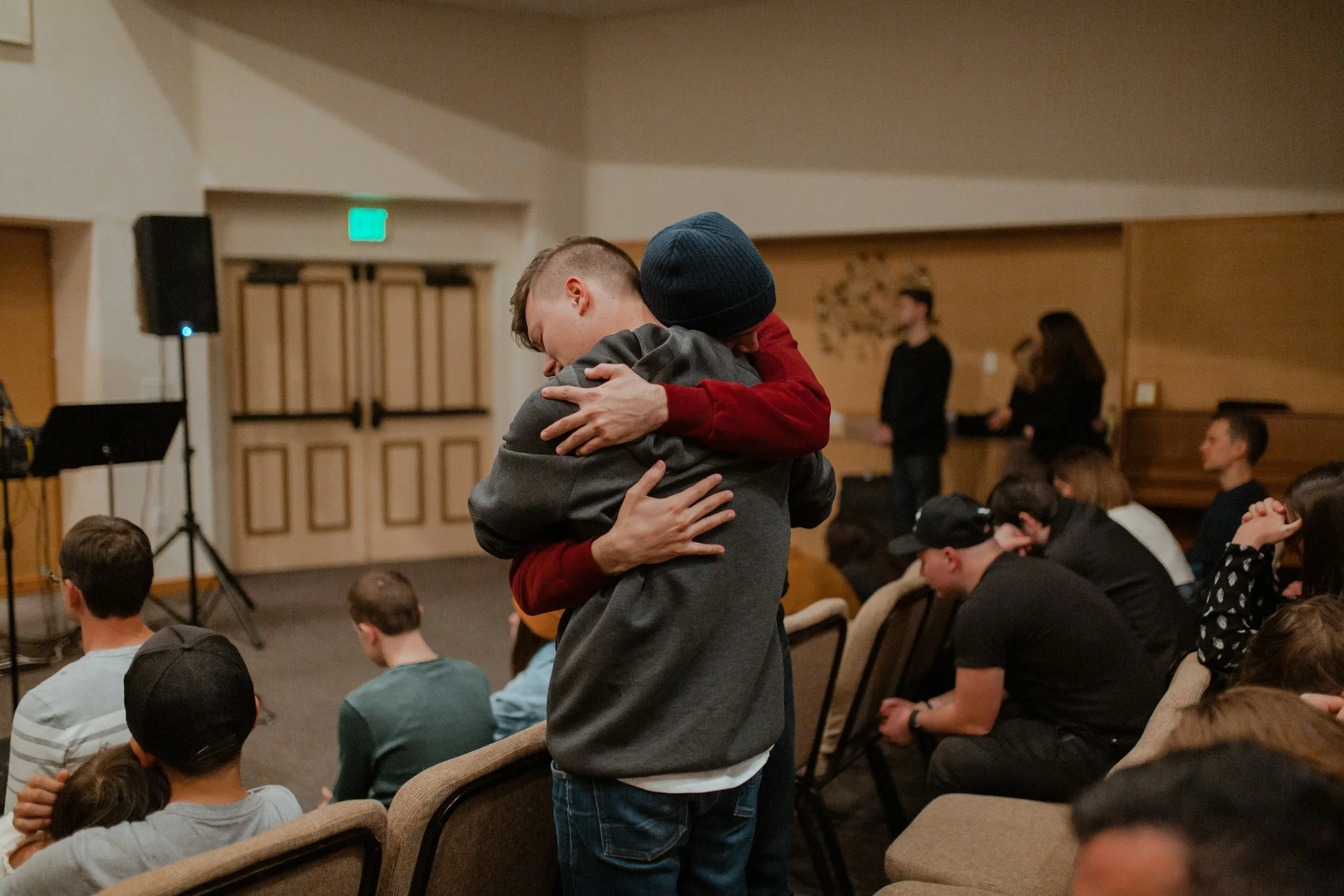Two people sharing a hug in an indoor setting with an audience seated, some with heads bowed or eyes closed, and a man standing in the background near a piano.