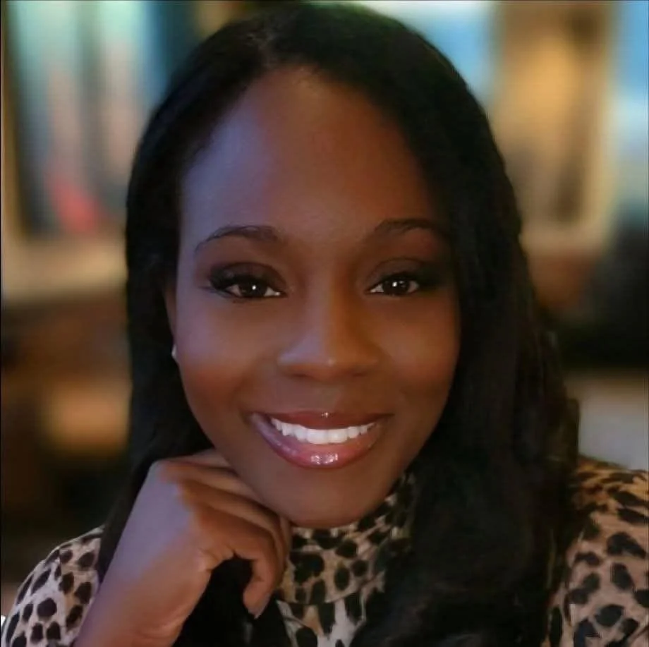 A smiling woman with dark hair in loose waves, wearing leopard print clothing, resting her chin on her hand in a warmly lit setting.