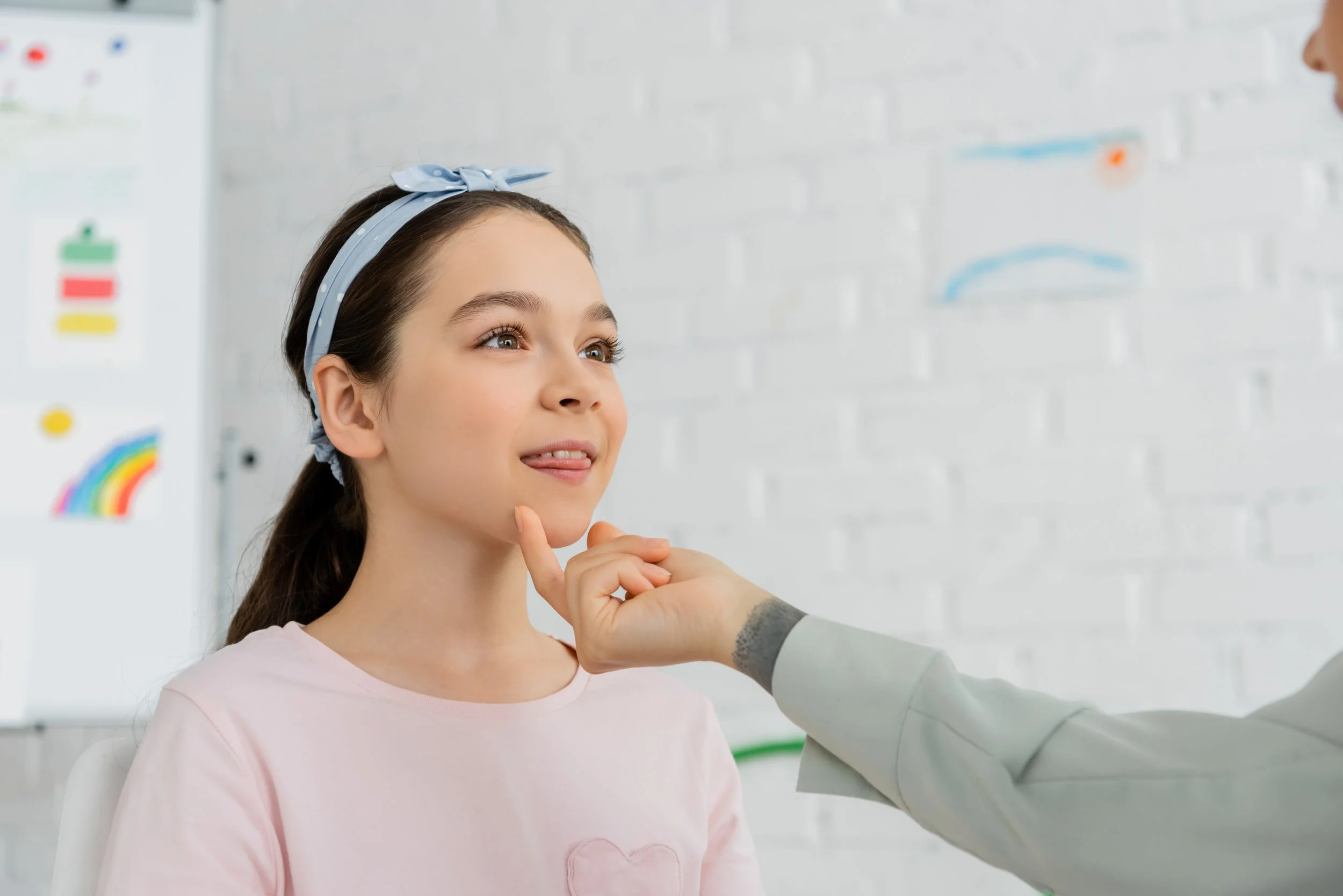 A young girl with brown hair, wearing a pink shirt and a blue headband, looks upward while her speech therapist works with her on her articulation disorder.