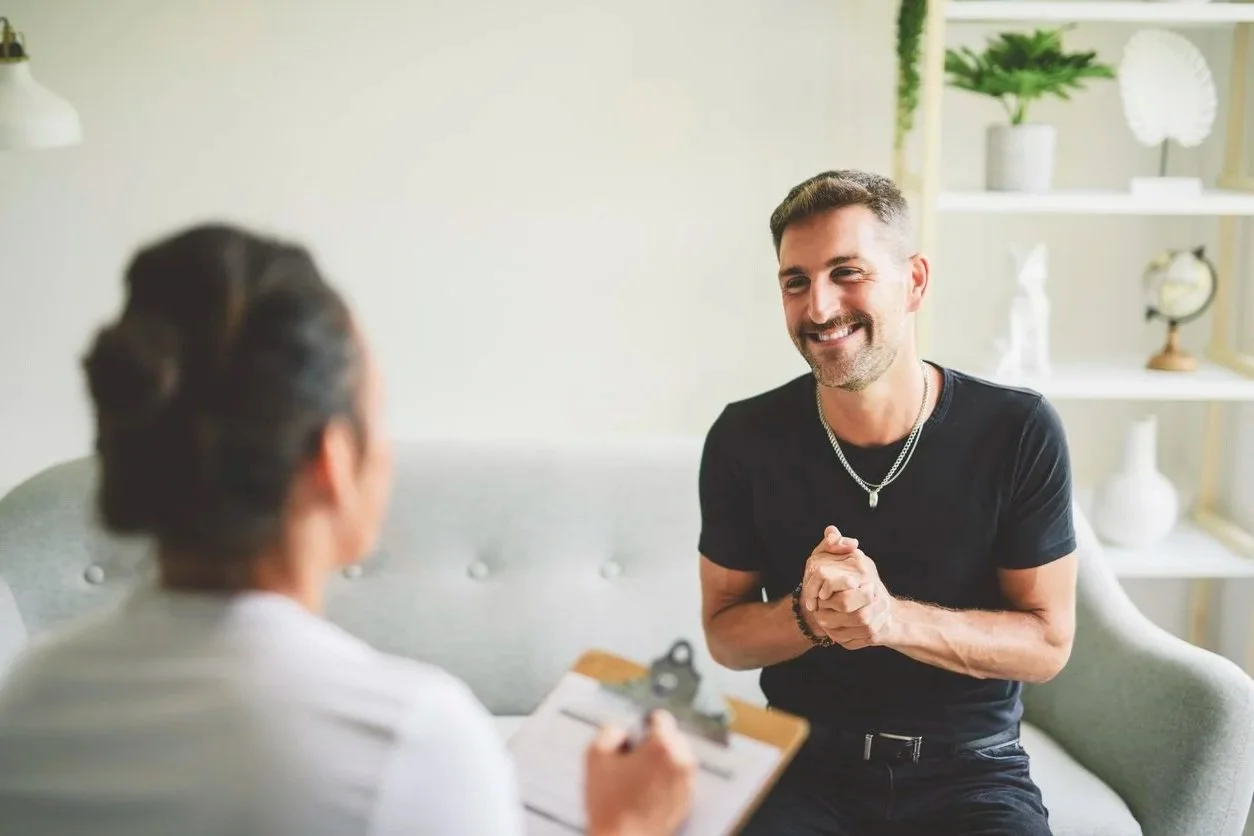 A man smiling and sitting on a sofa, talking to his speech therapist in Milwaukee, WI, during his evaluation, in a bright room with white shelves and plants in the background.