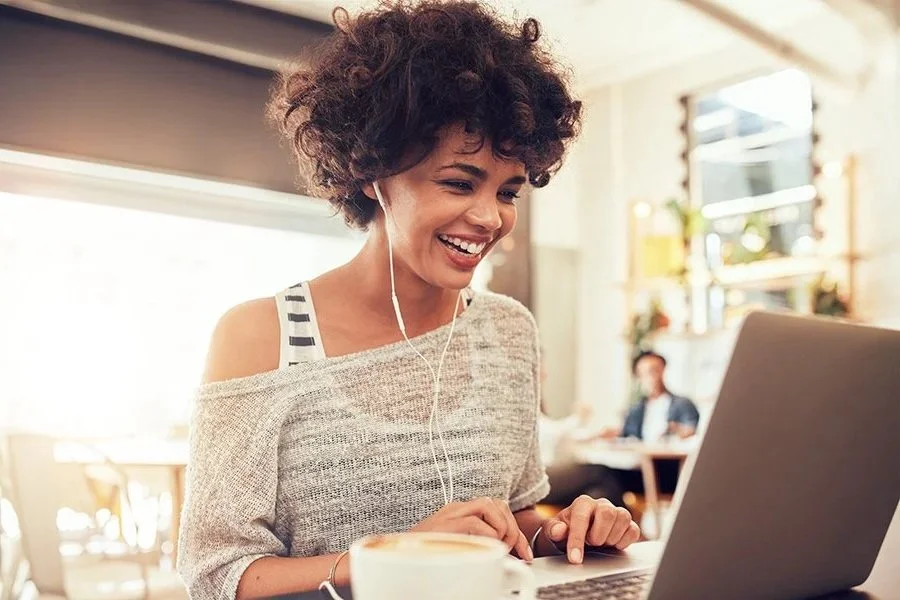A woman smiling and enjoying herself as she uses a laptop in a bright, modern cafe, wearing earphones and a casual off-shoulder top.