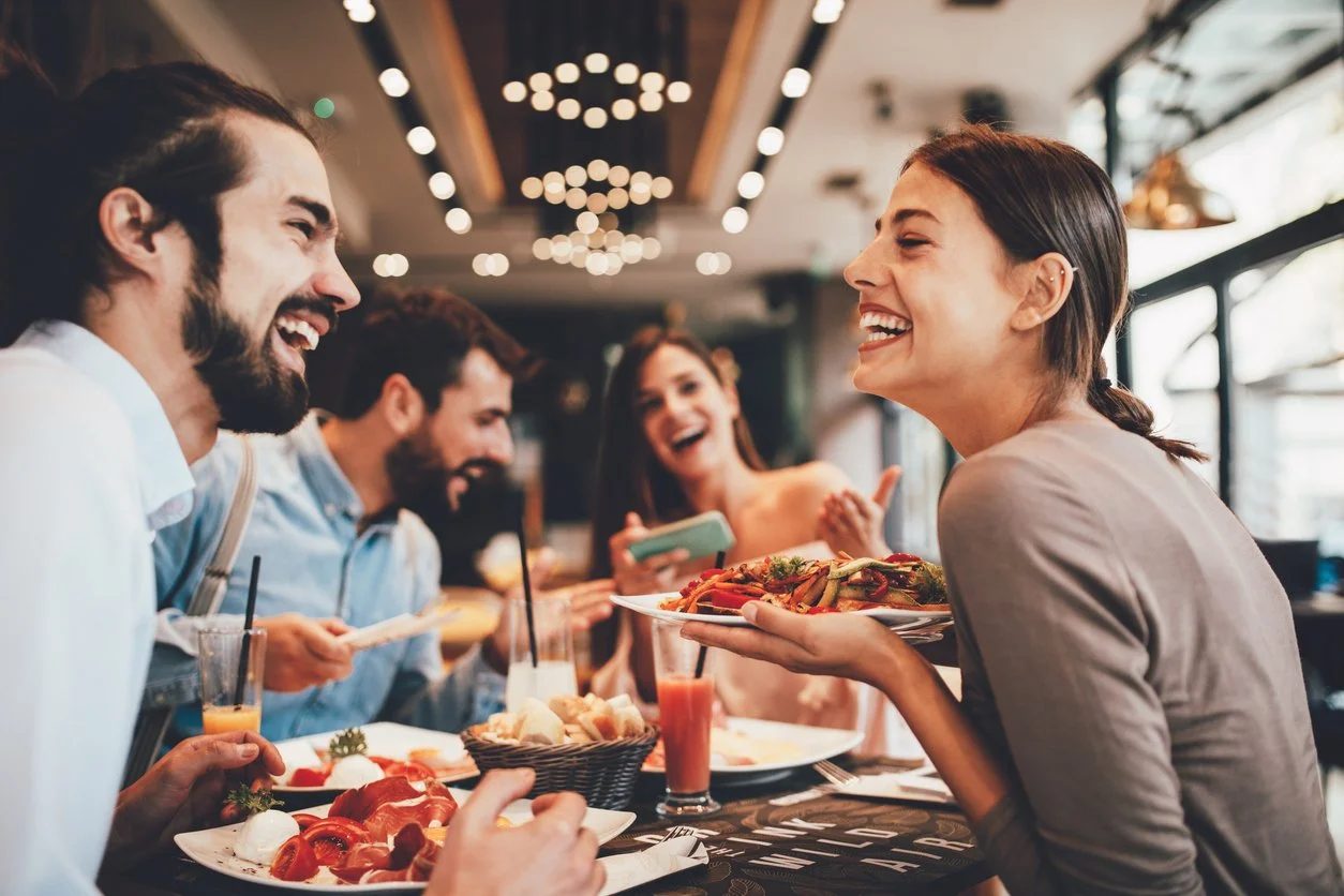 A group of friends enjoying a meal together at a restaurant, with two women and three men smiling and laughing.