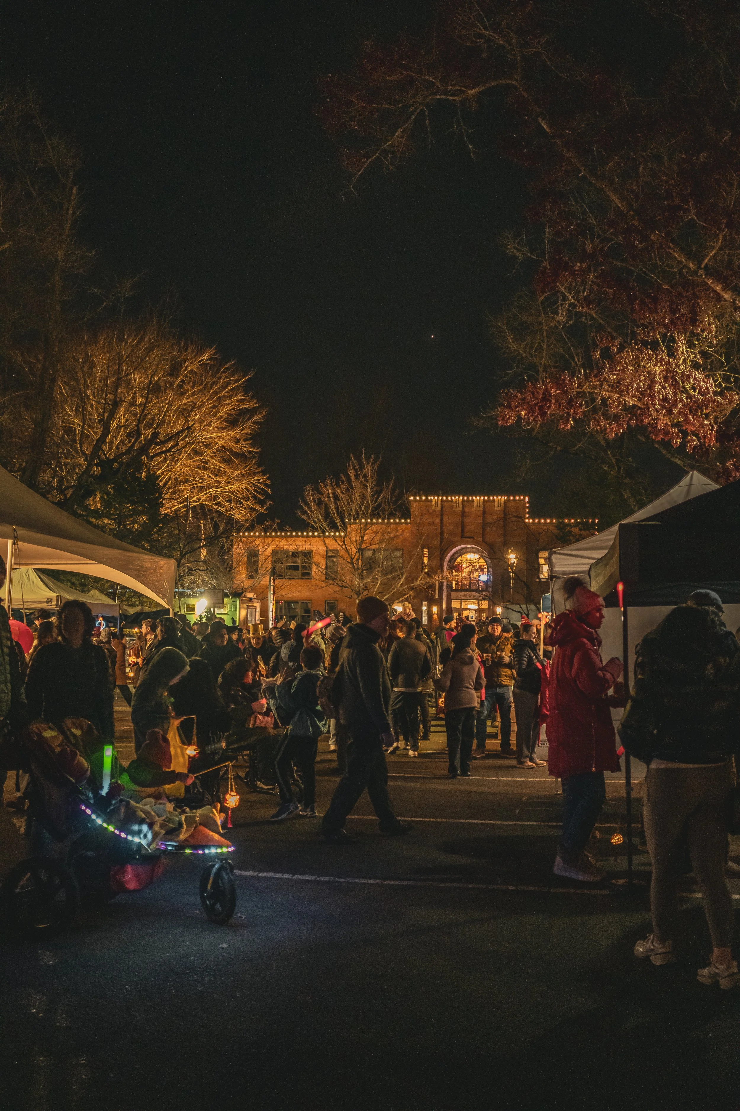 Night scene of a crowded outdoor event with tents, people walking, socializing, and children in strollers, with a brick building decorated with string lights in the background.