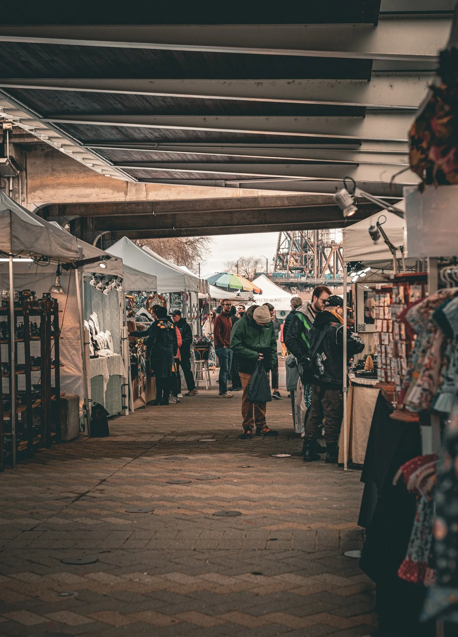 People shopping at an outdoor market under a bridge, with vendor tents and booths selling various goods, some with umbrellas and displays, during daytime.